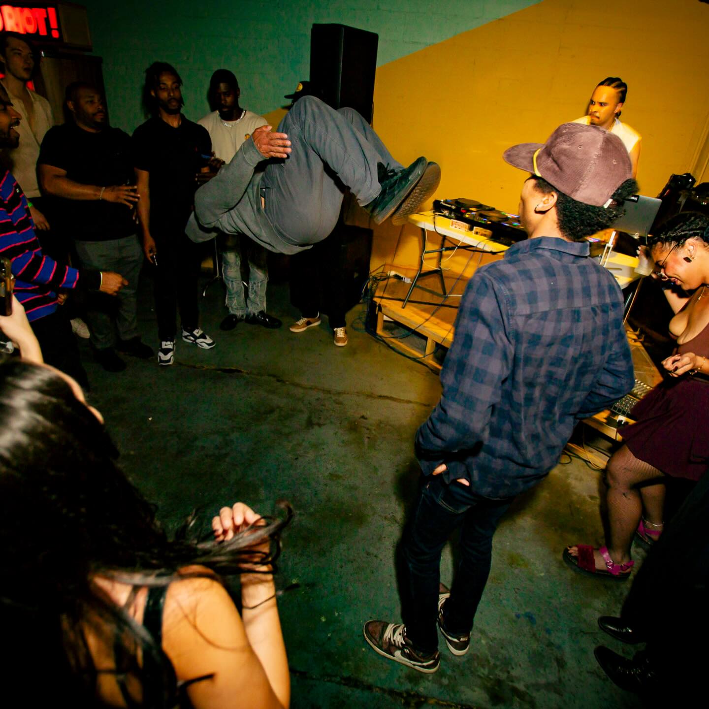 Person performing a backflip in a room with a group of people watching, DJ equipment on a table, colorful walls.