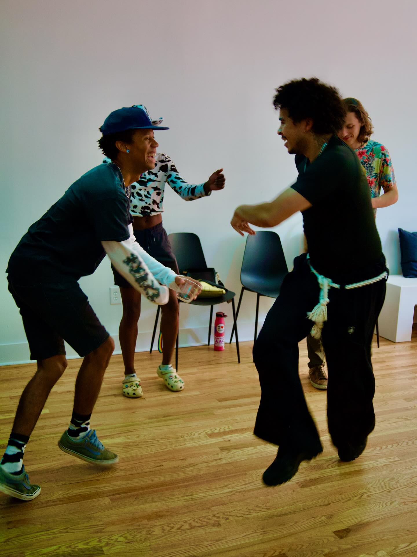 Four people dancing indoors, two men and two women, with chairs in the background, wooden floor, and plain wall.