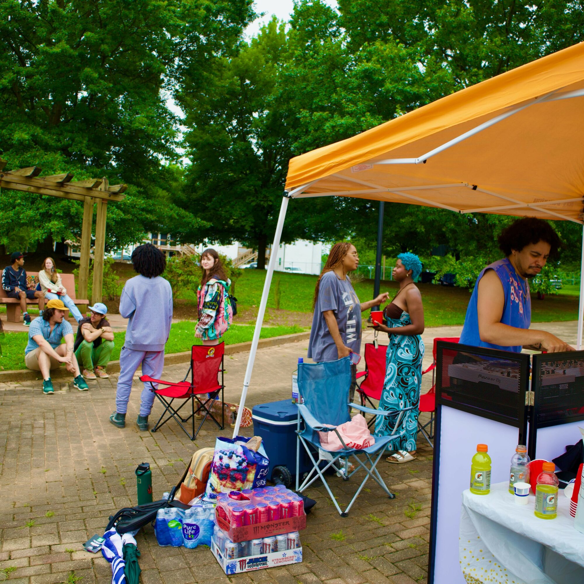 People gather outdoors under a yellow canopy, with children sitting on the ground and others standing near a grill, in a park setting.