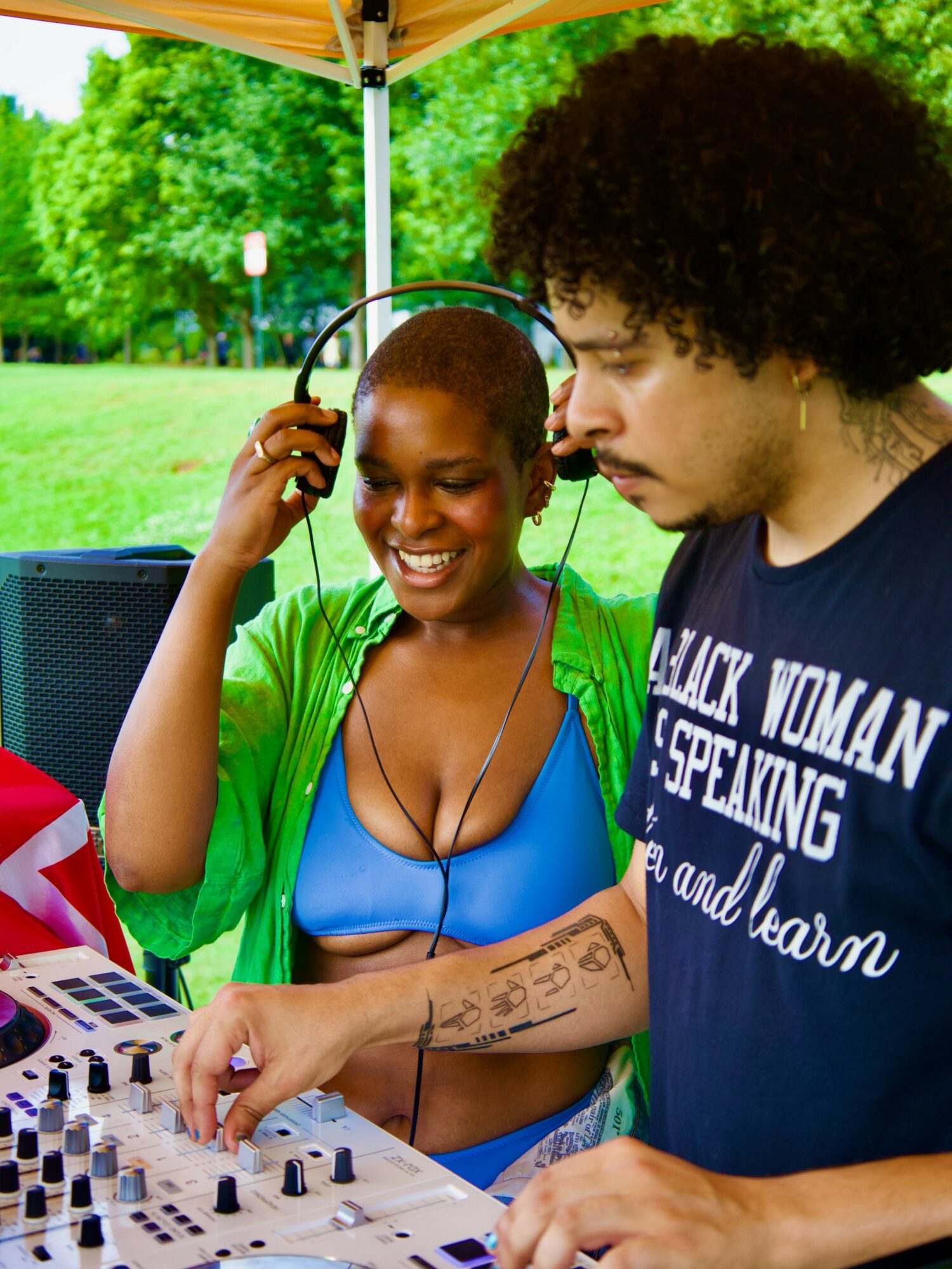 Two people, one smiling and wearing headphones, are at a DJ setup outdoors with green trees in the background.