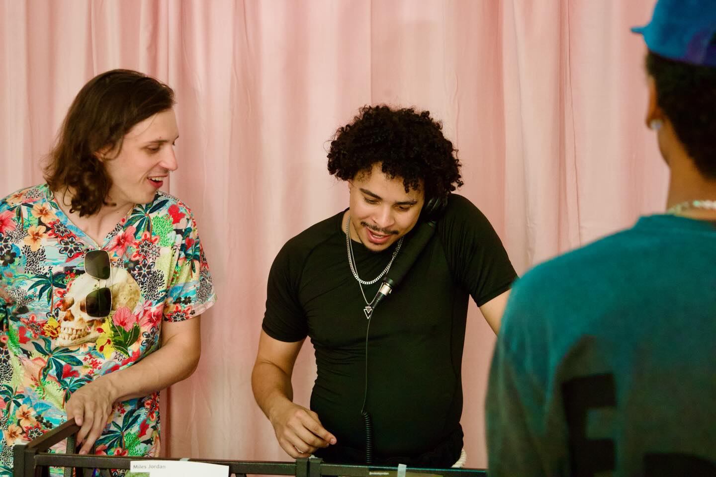 Three people smiling and talking indoors with pink curtains in the background.