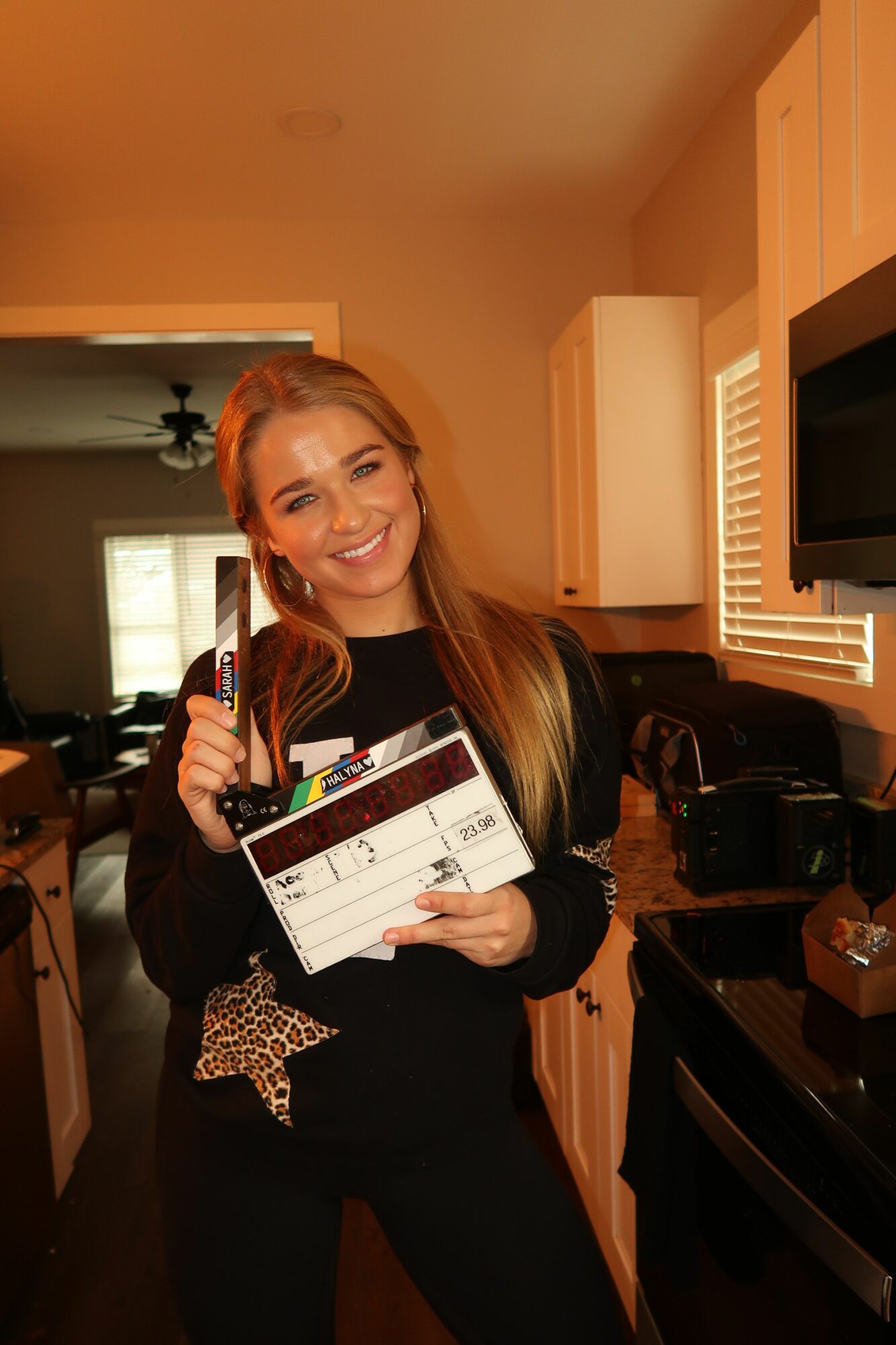 Woman with long blonde hair holding a clapperboard and smiling indoors, with a window and furniture in background.