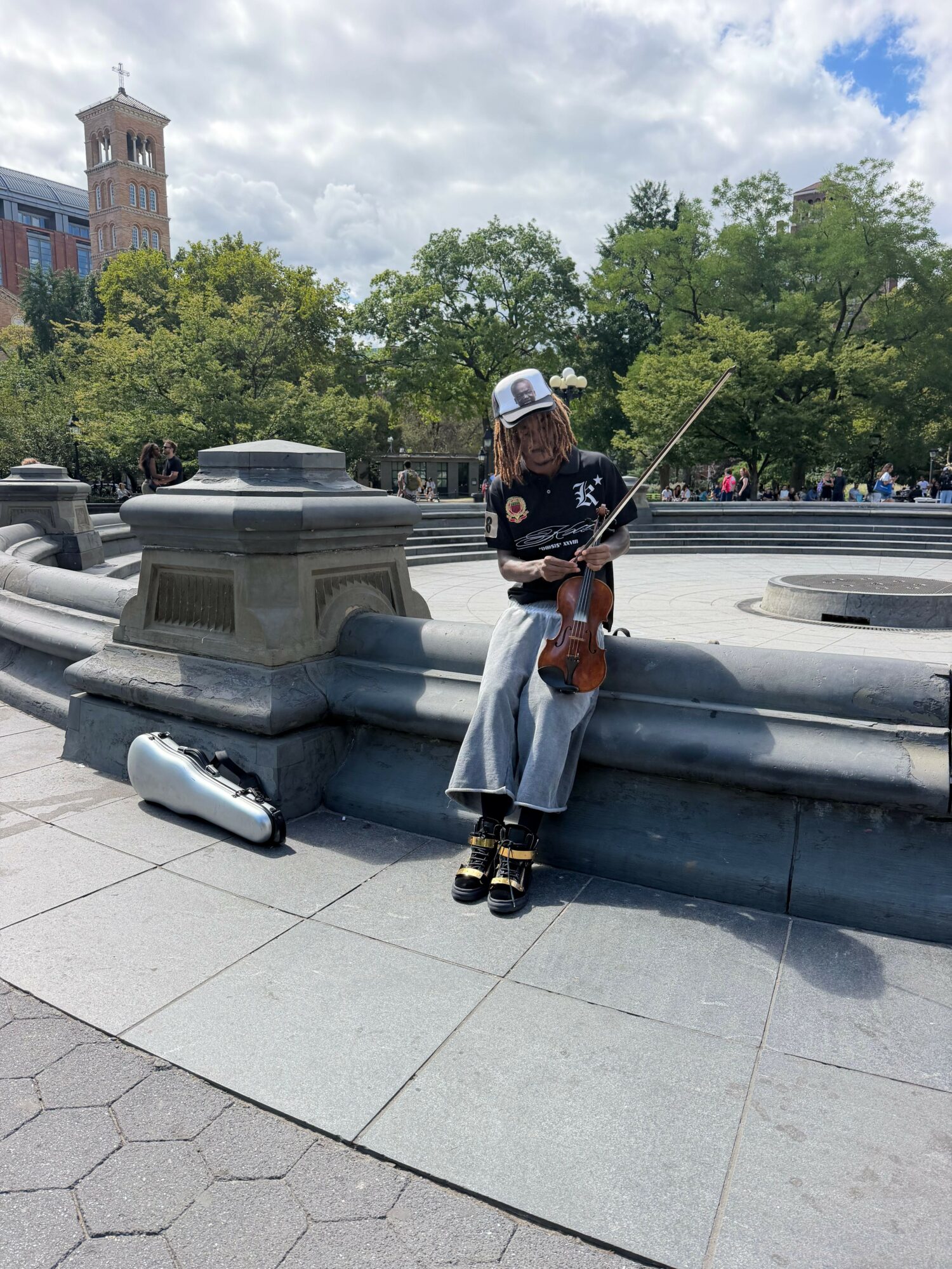 Person fishing with a rod near a fountain in a park, trees and a building in the background.