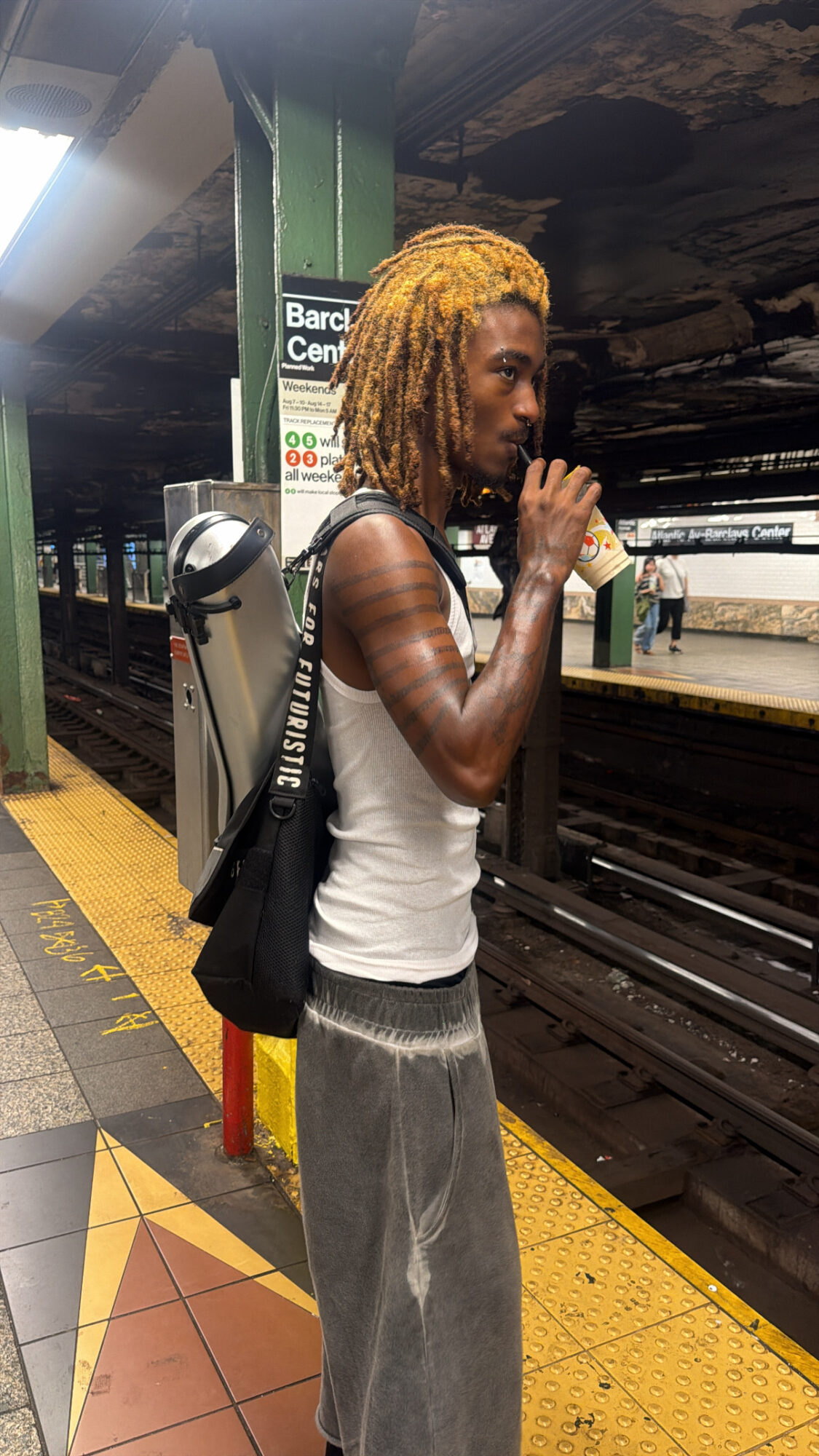 Person with dreadlocks standing on subway platform, holding a phone, wearing a white tank top and gray pants.