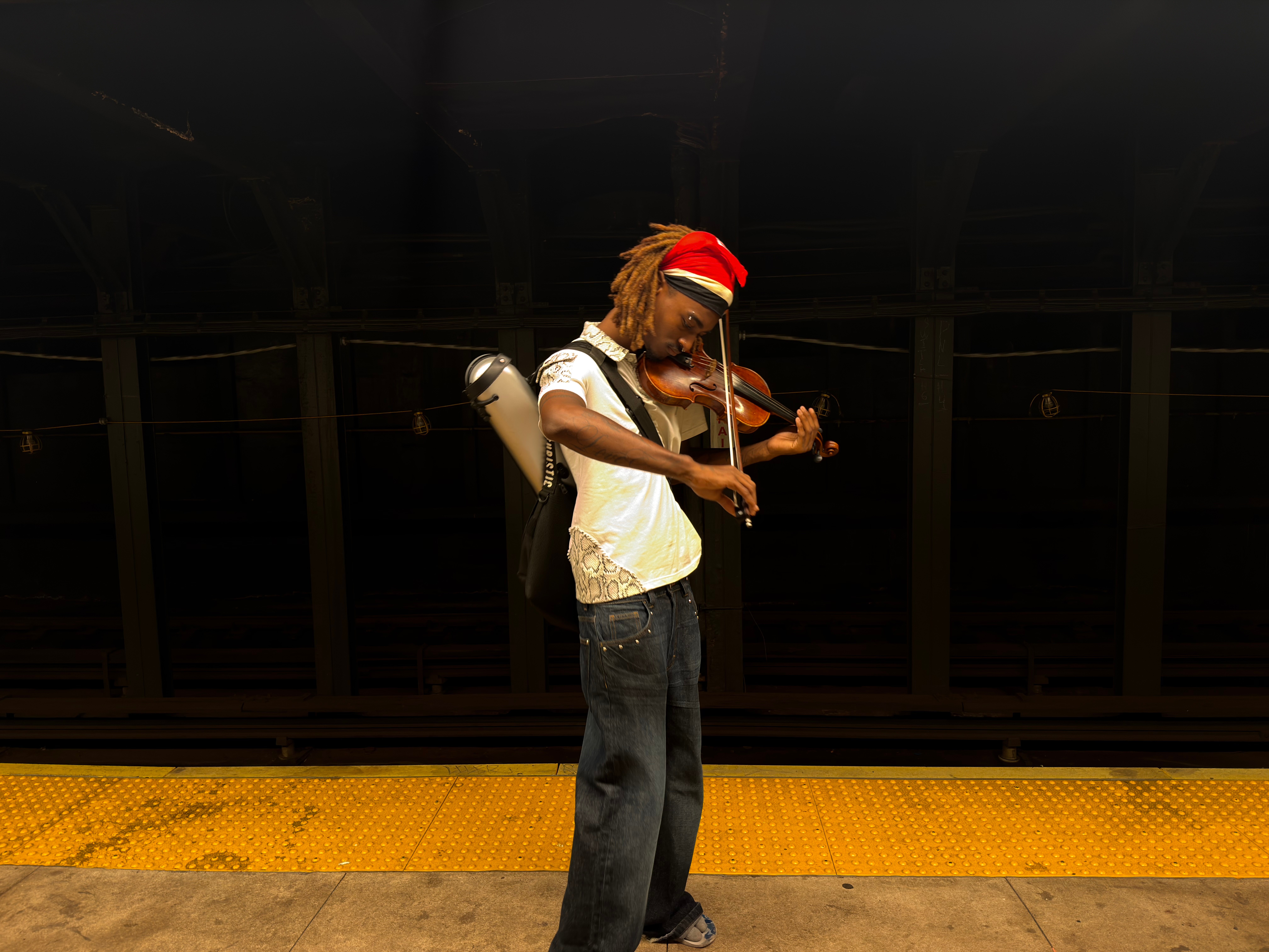 Person with dreadlocks wearing a red headband playing a violin on stage, with a dark background and yellow floor strip.