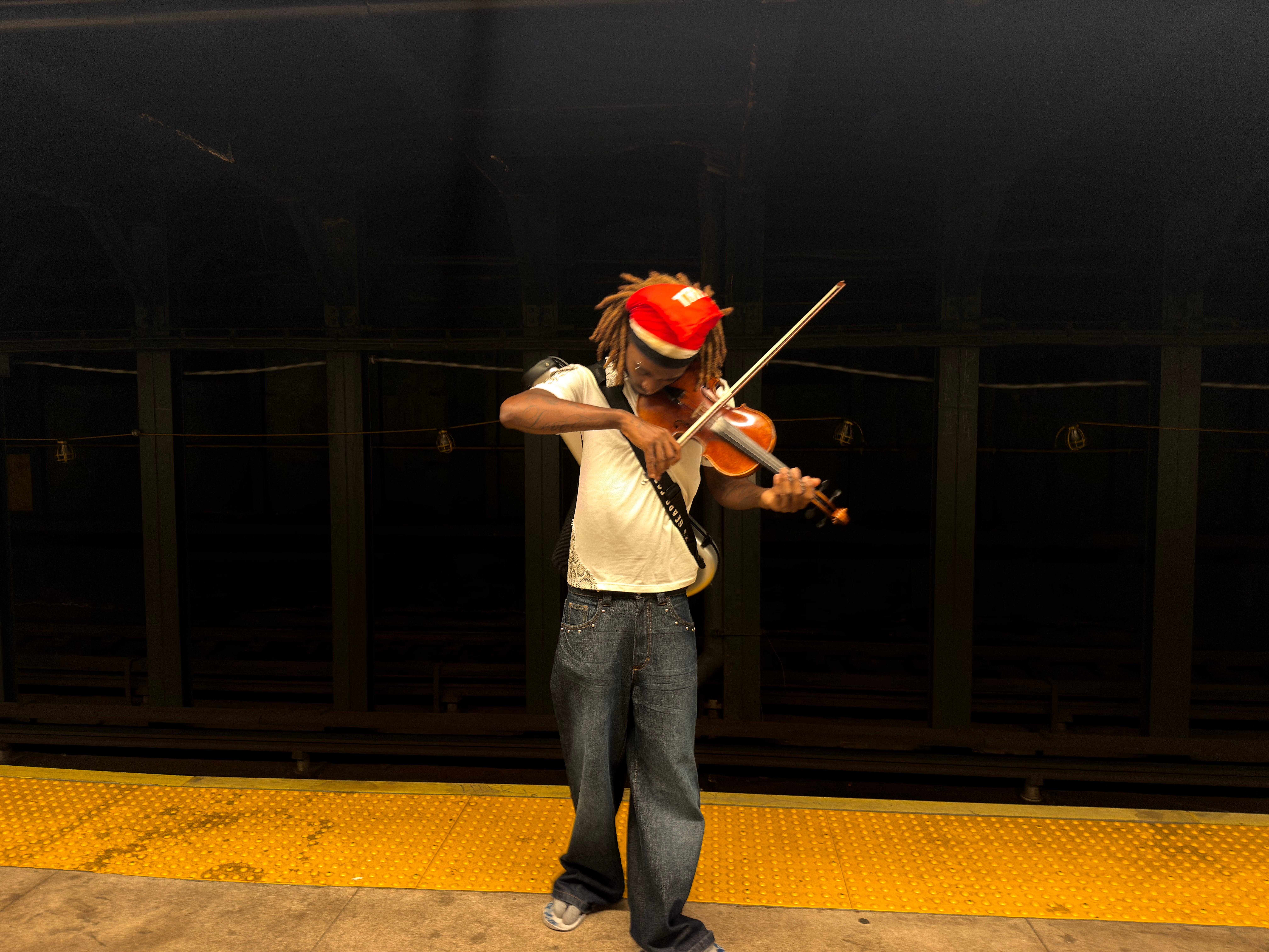 Person playing violin on subway platform, wearing a red hat, white shirt, and jeans, with dark background and yellow safety strip.