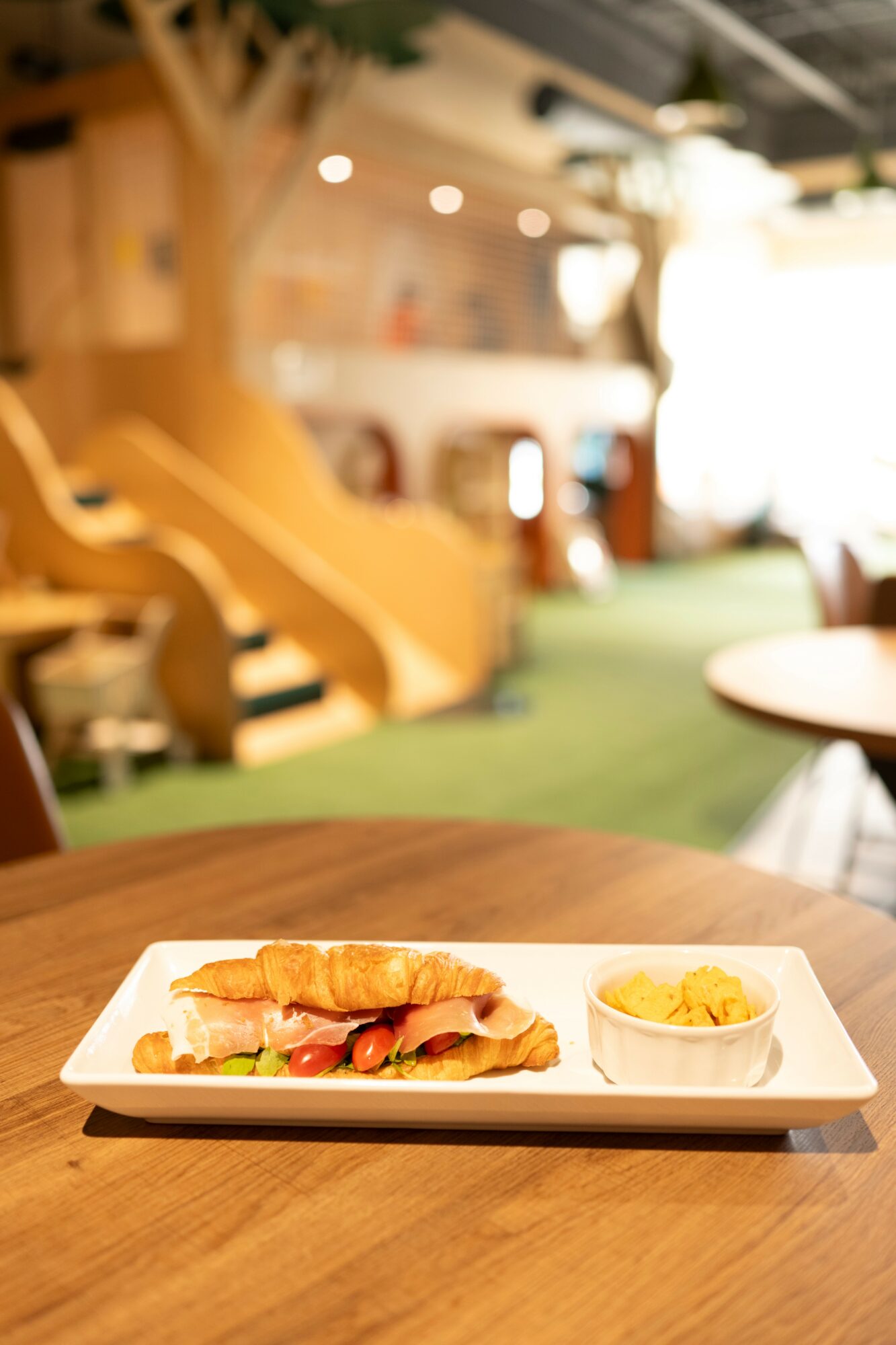 A plate with a sandwich and a side dish on a wooden table in a blurred indoor setting.