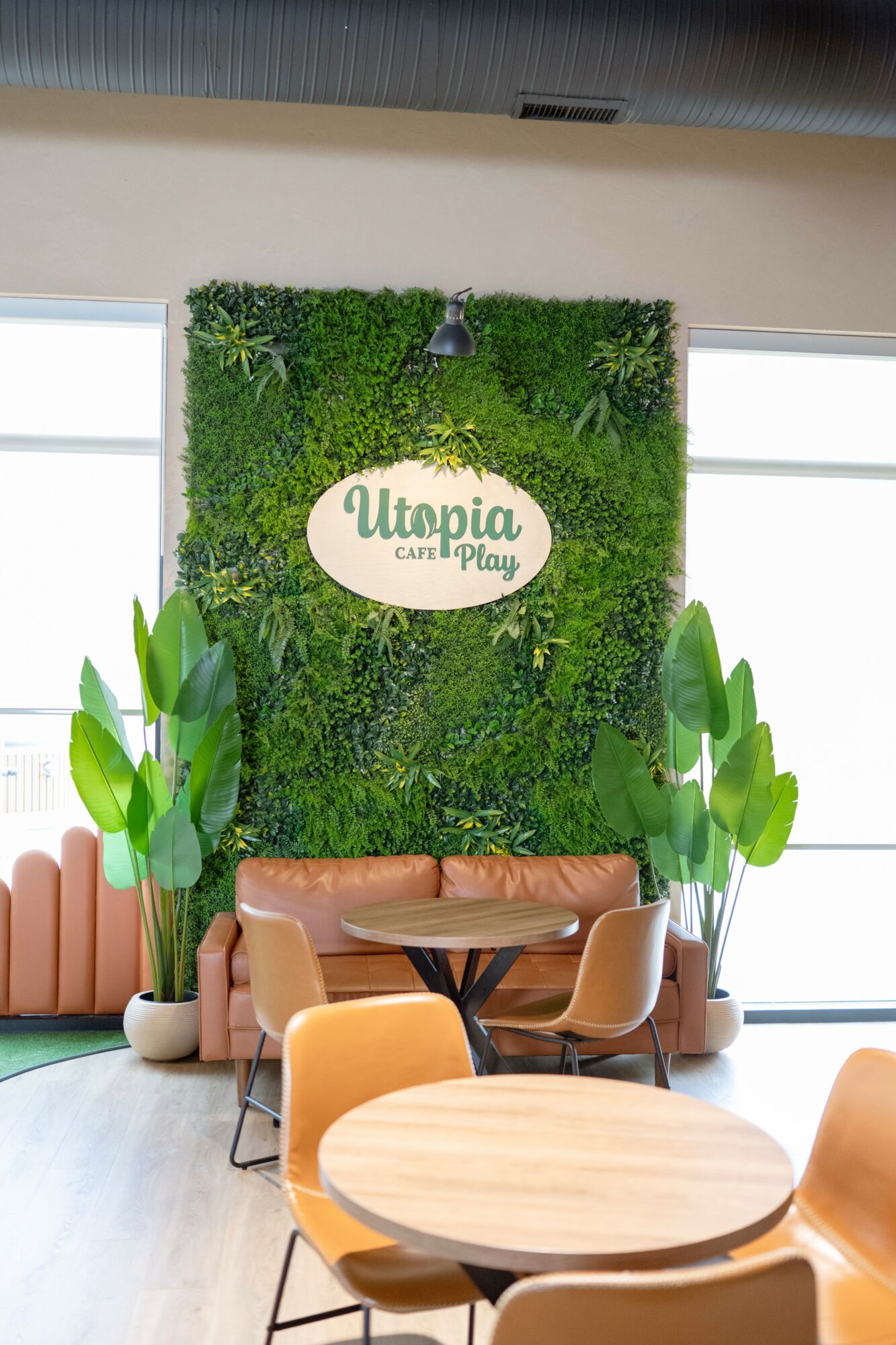 Indoor seating area with a green wall, a sign reading 'Utopia Cafe & Play,' and plants, with tables and chairs.