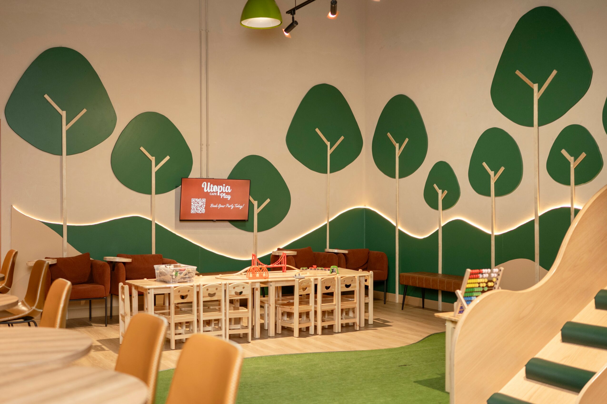 Indoor space with green tree and hill wall mural, wooden furniture, and a table with white chairs, children’s play area.