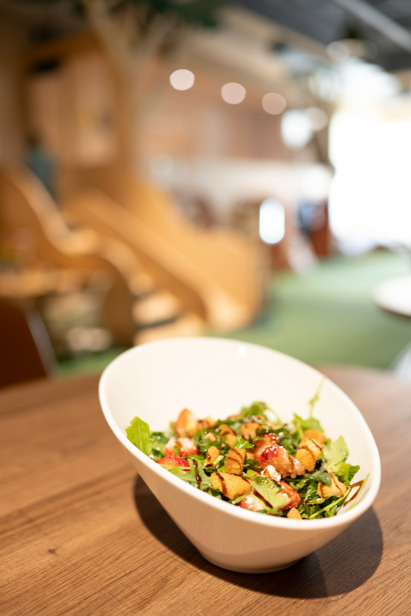 Bowl of salad with greens, tomatoes, and other vegetables on a wooden table, blurred background.