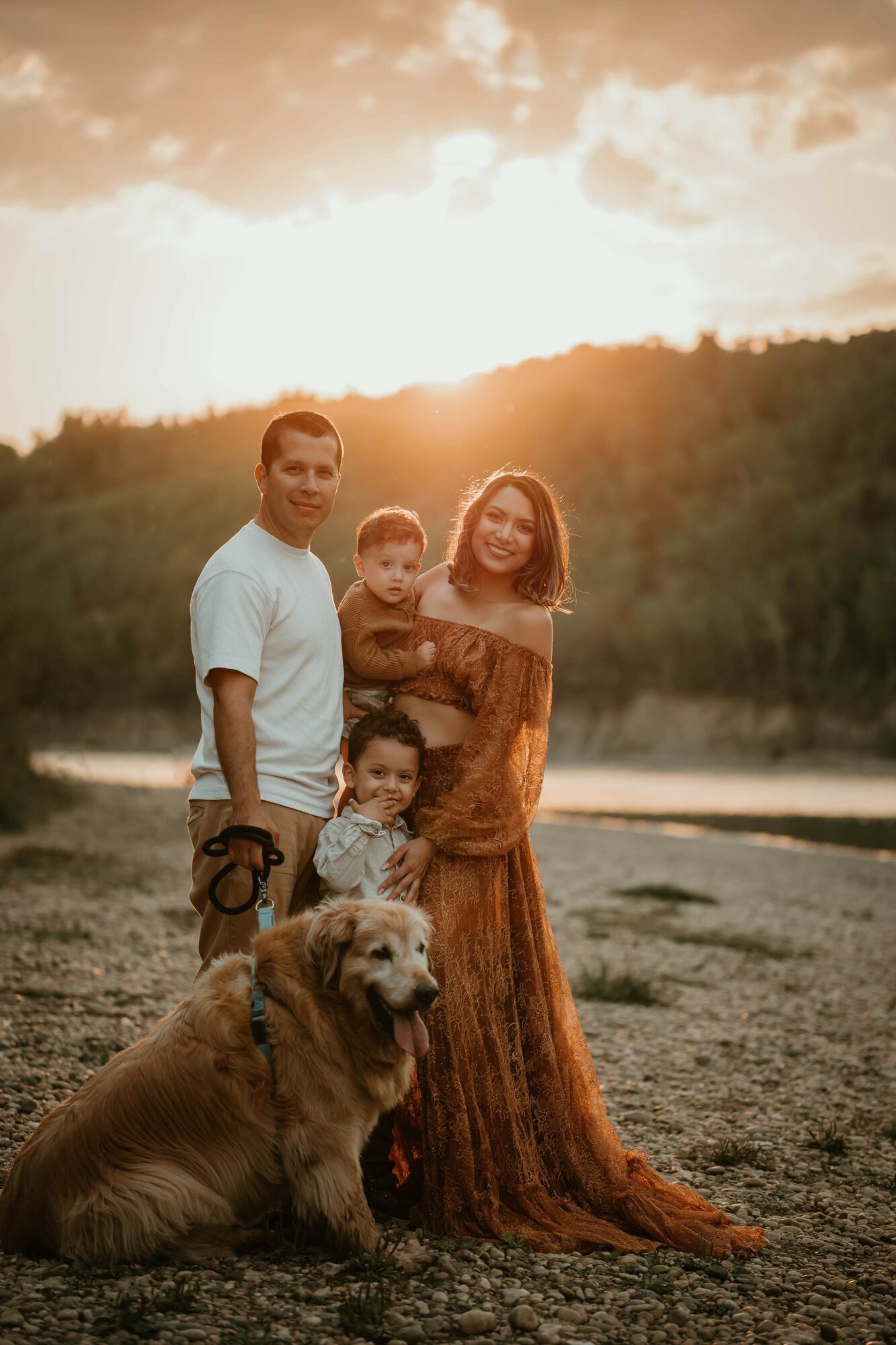 Family of four with a dog outdoors at sunset, standing on a sandy area near water, smiling, with trees in background.