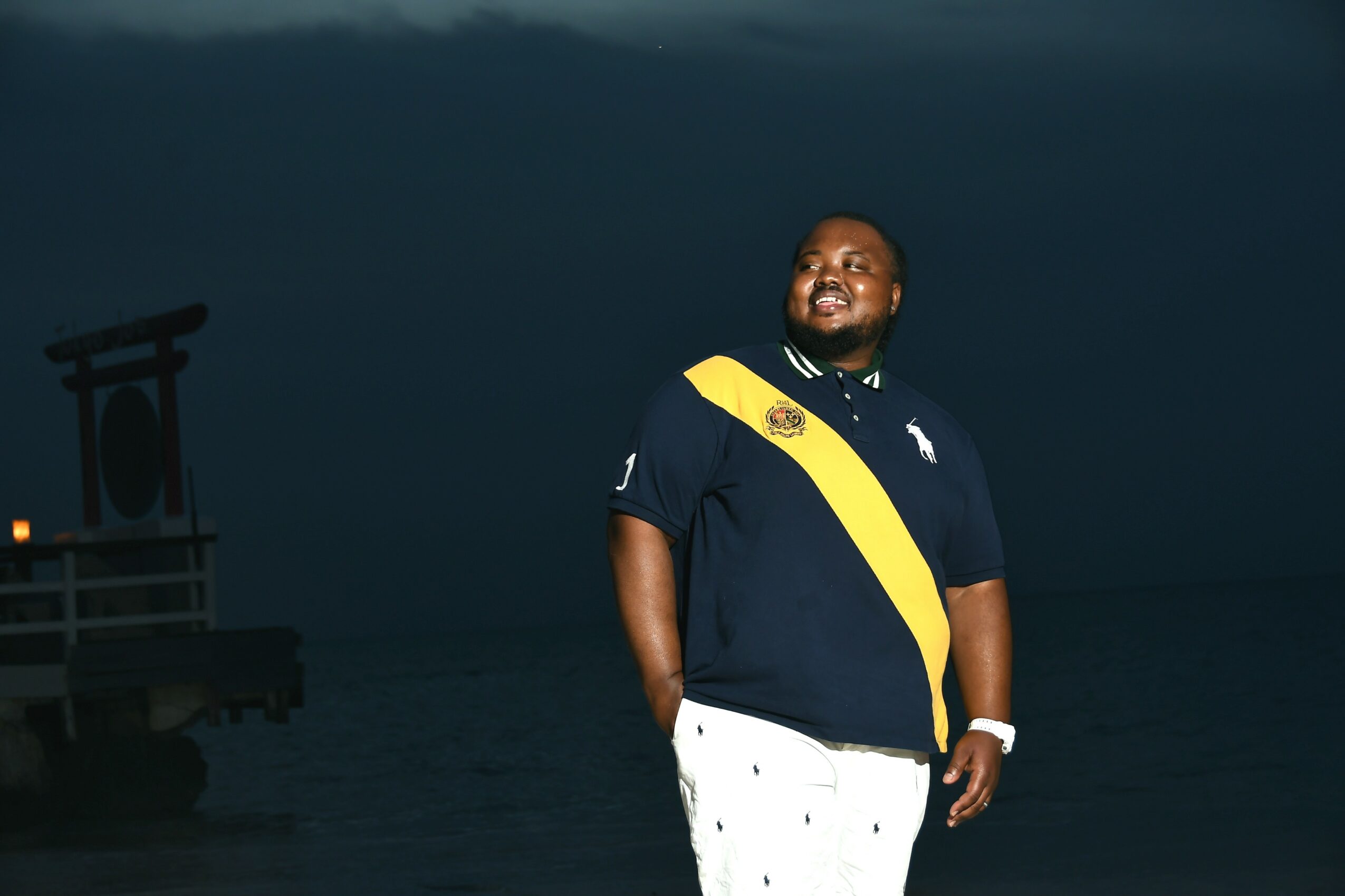 Person standing outdoors at night, smiling, wearing a dark shirt with yellow stripe, white pants, with a structure in the background.