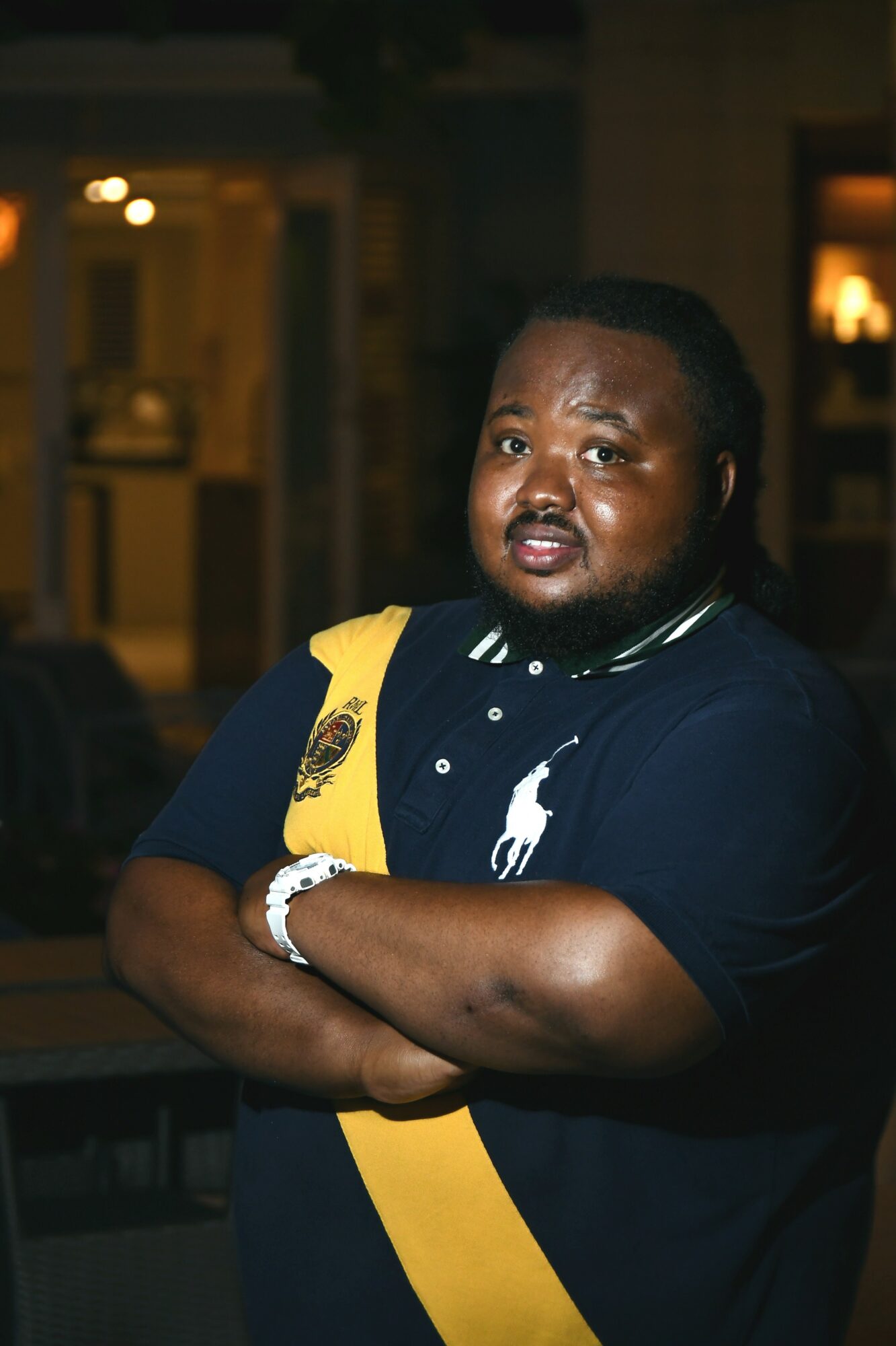 Man with crossed arms wearing a navy polo shirt with a yellow strap, indoors at night, looking at the camera.