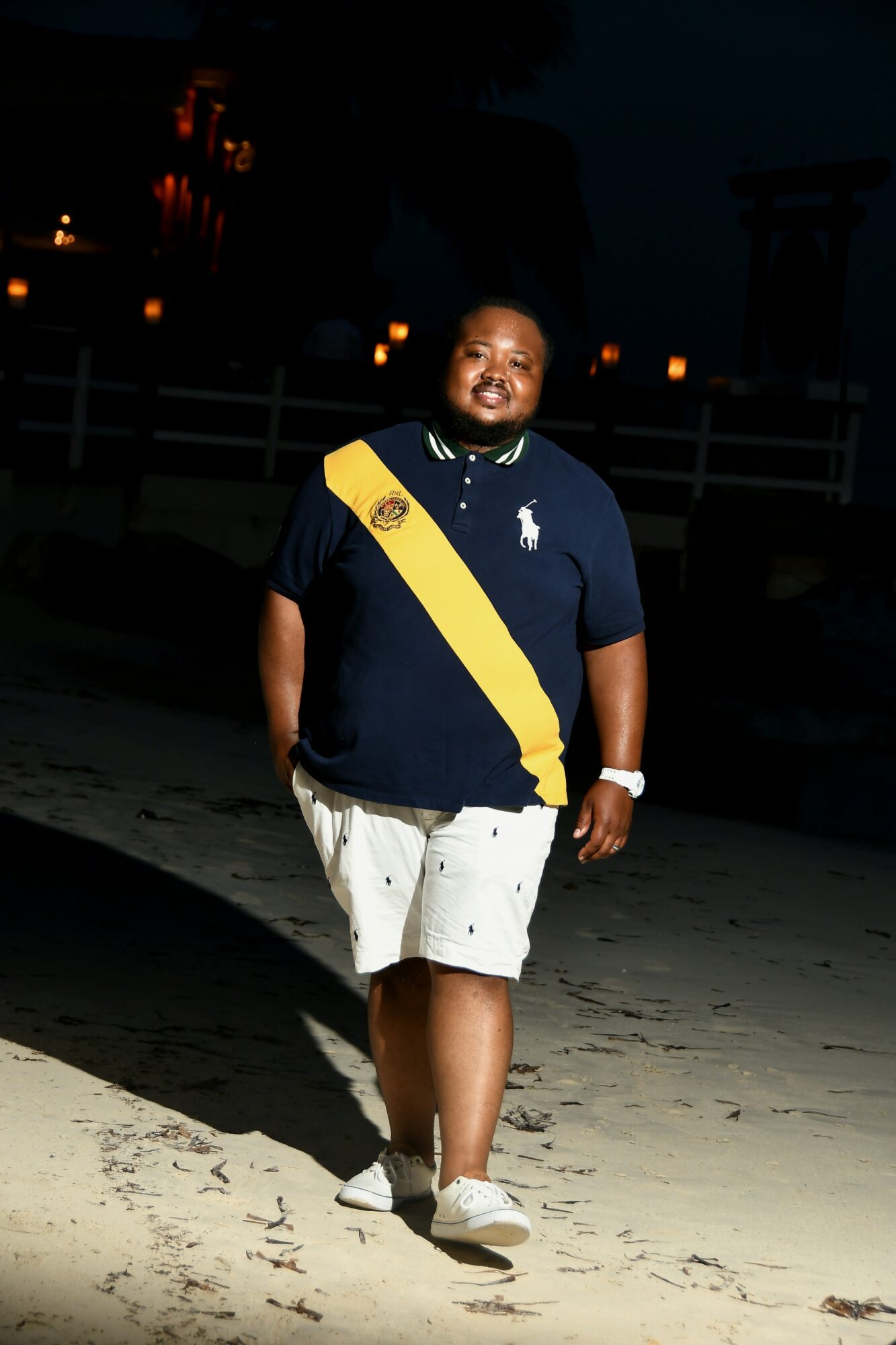 Person walking outdoors at night, wearing a dark shirt with a yellow sash, white shorts, and sneakers, smiling.