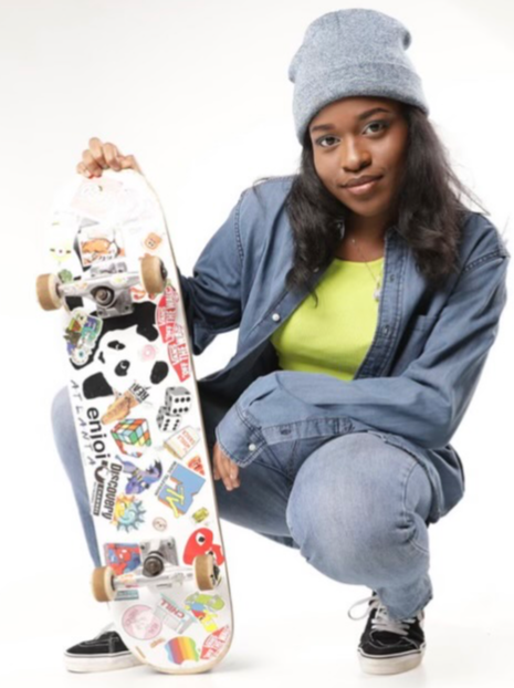 Young woman crouching with skateboard, wearing gray beanie, denim jacket, and lime green shirt, against white background.