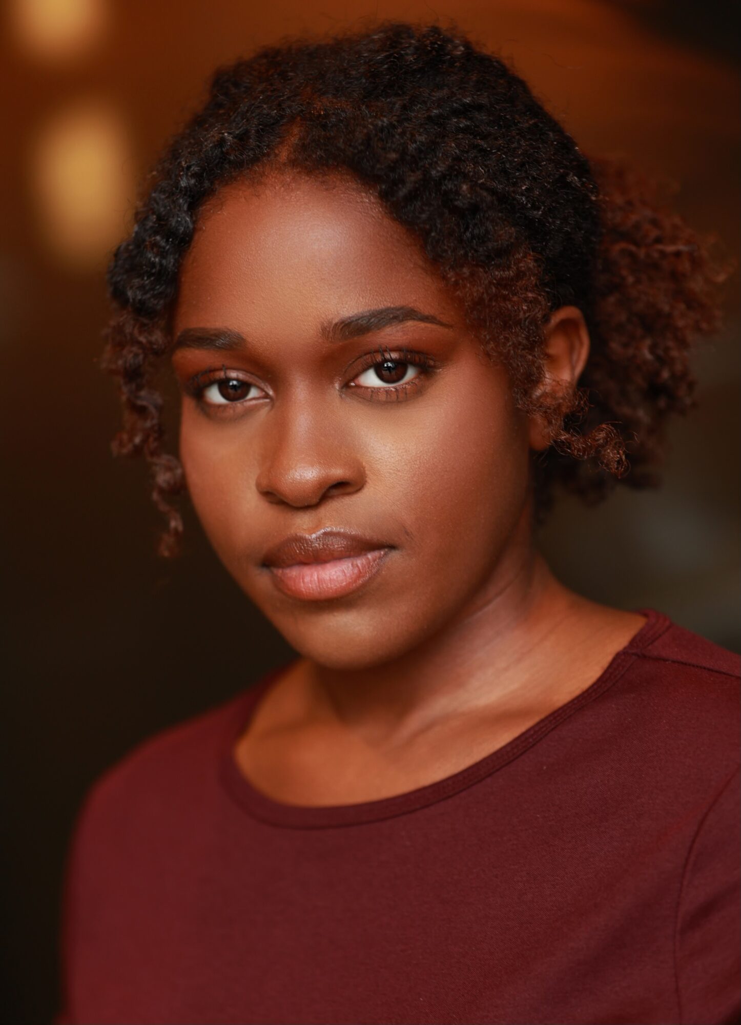 Portrait of a young woman with natural curly hair, wearing a maroon top, looking directly at the camera.