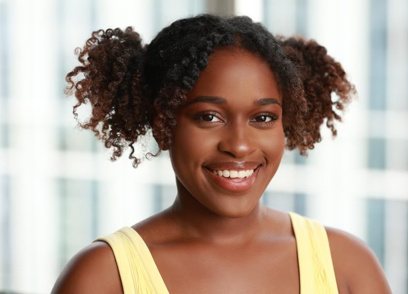 Smiling woman with curly hair in puffs, wearing a yellow top, standing indoors with a window background.