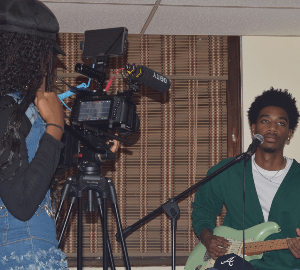 Person with curly hair filming a young man with an electric guitar and microphone, in a room with wooden blinds.