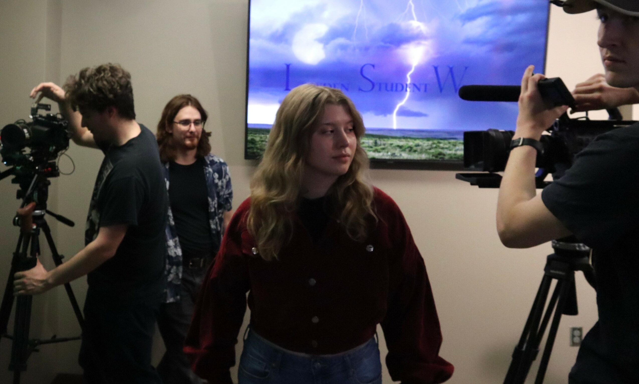 Group of people, including a woman with long hair, in a room with cameras and a large screen displaying a cloudy sky.