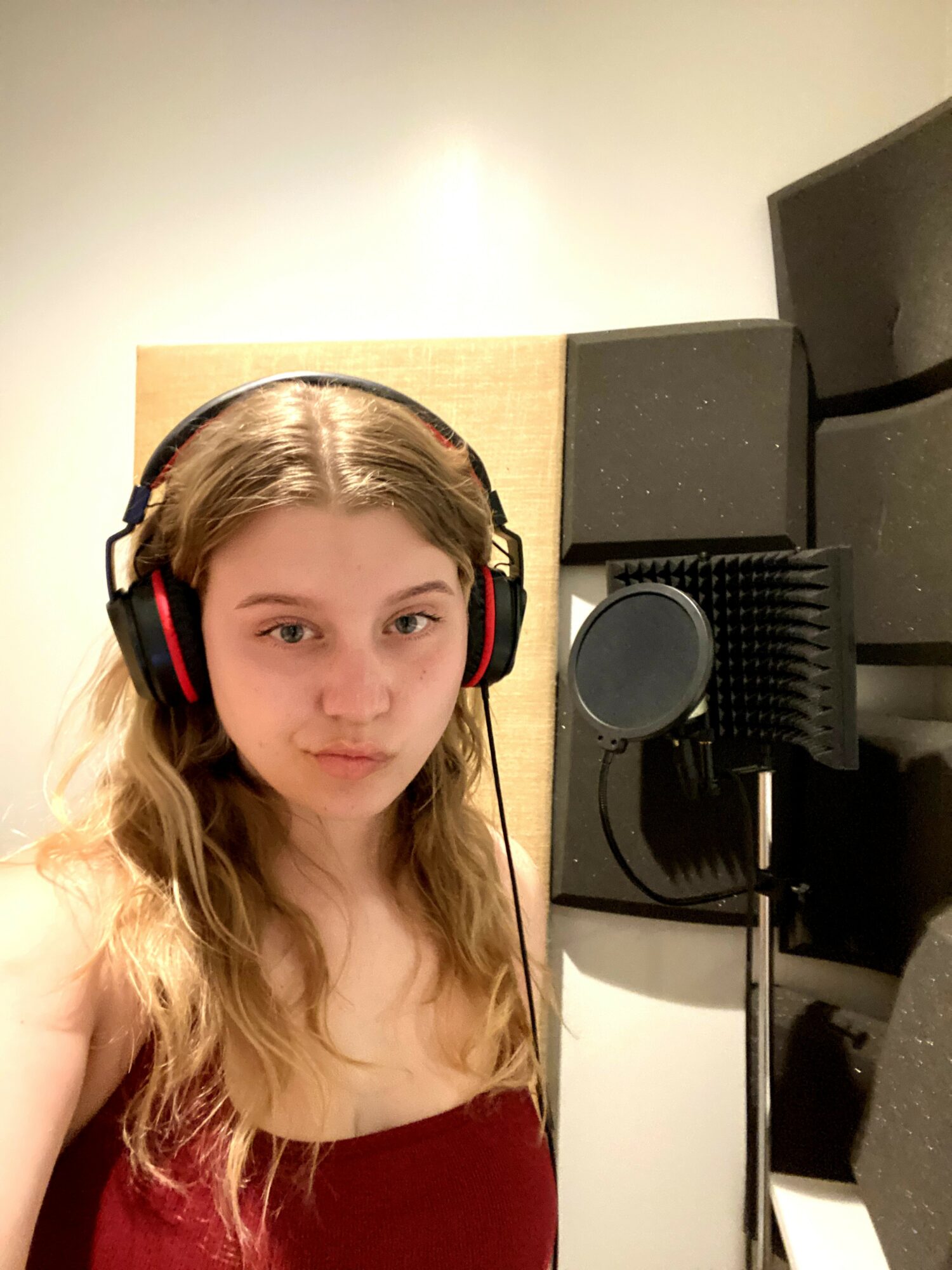 Young girl with long hair wearing headphones in a recording studio, standing near a microphone and soundproof panels.