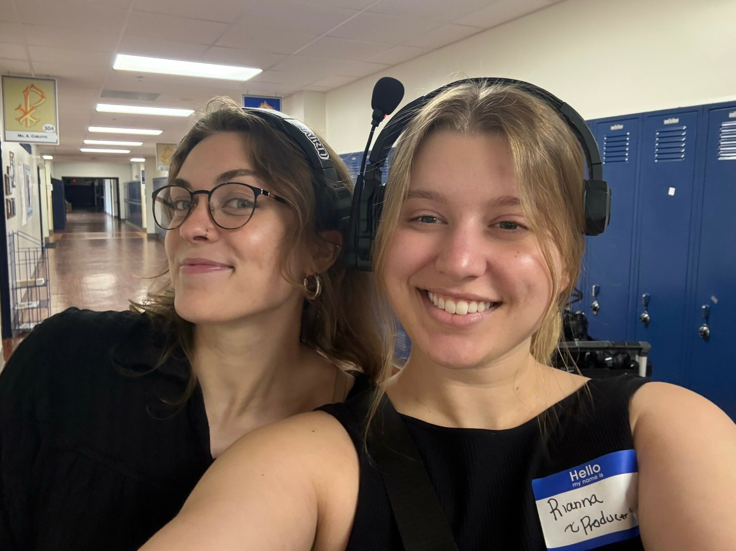 Two young women smiling, one wearing glasses and headphones, in a school hallway with blue lockers behind them.