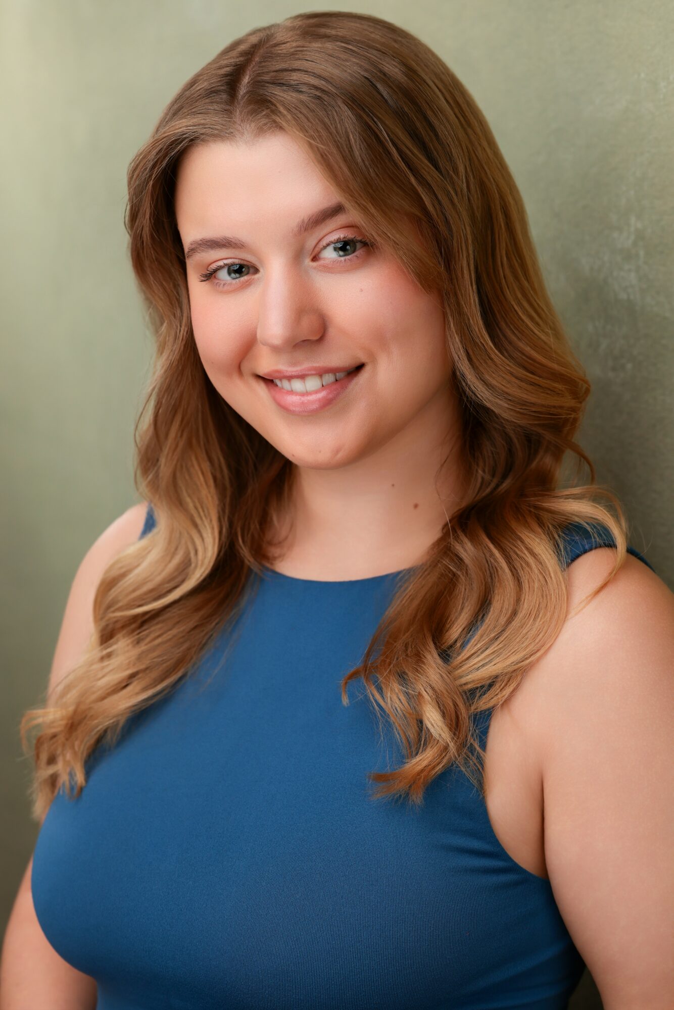 Young woman with long wavy hair smiling, wearing a blue sleeveless top, against a neutral background.