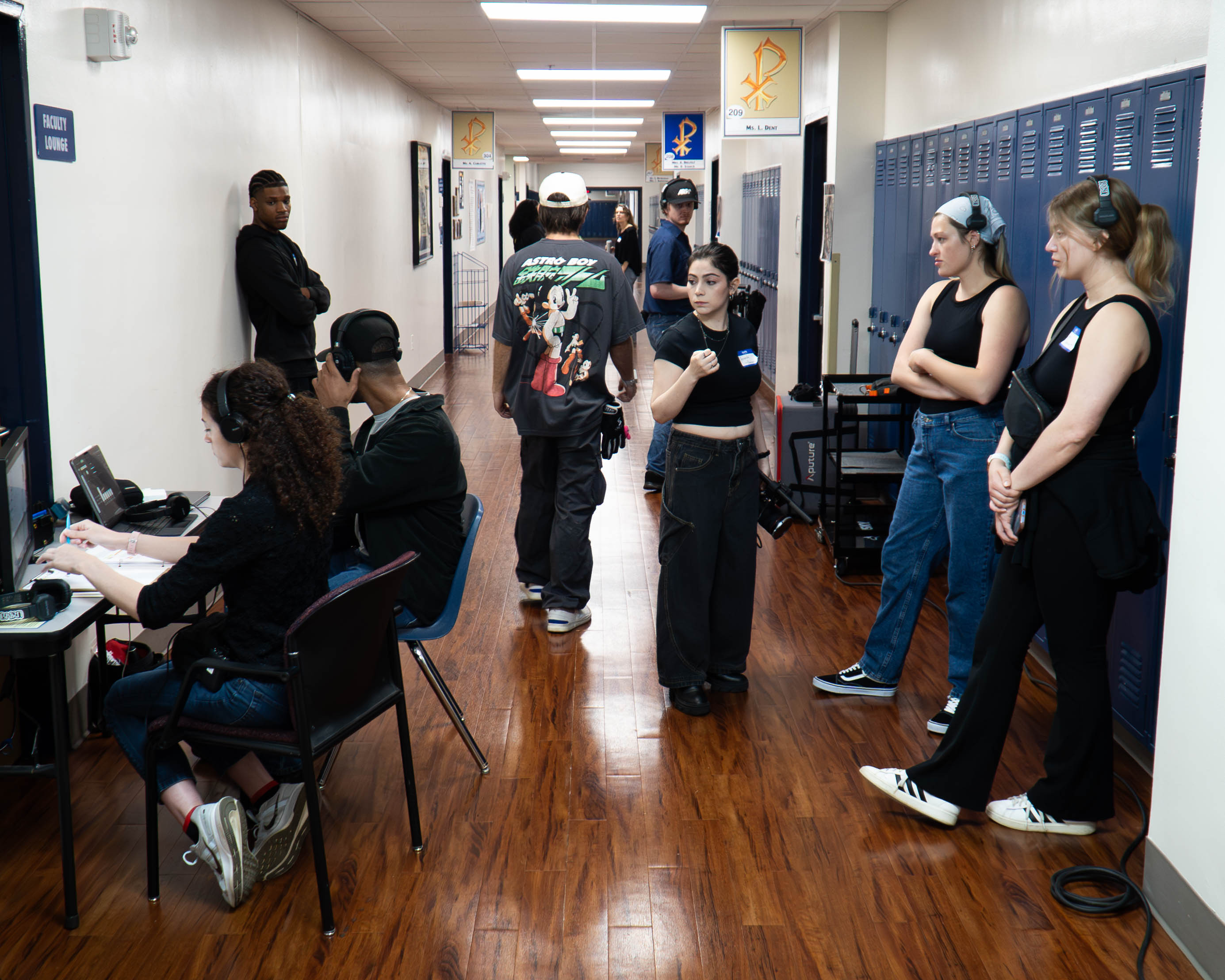 Students in a school hallway with lockers, some sitting at desks and others standing and talking, wooden floor, white walls.