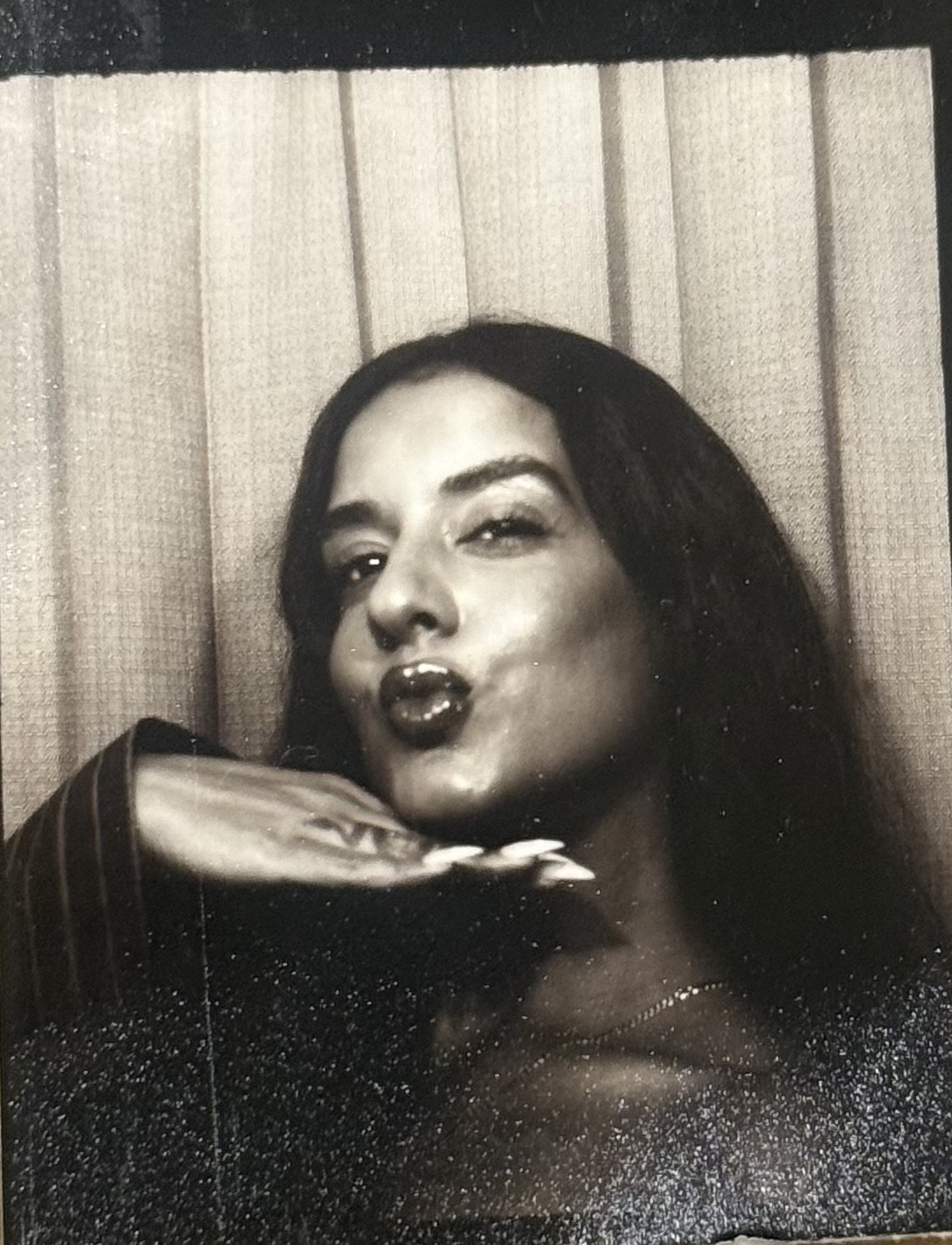 Woman with long dark hair making a kiss face, resting her chin on her hand, in front of a curtain.