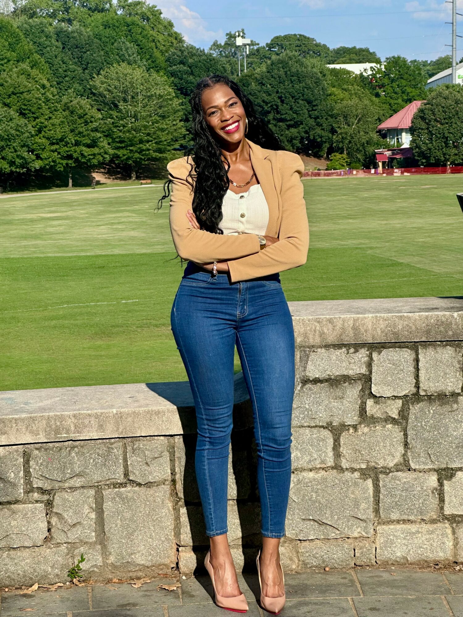 Smiling woman with long dark hair standing outdoors, arms crossed, in front of a stone wall and green park.