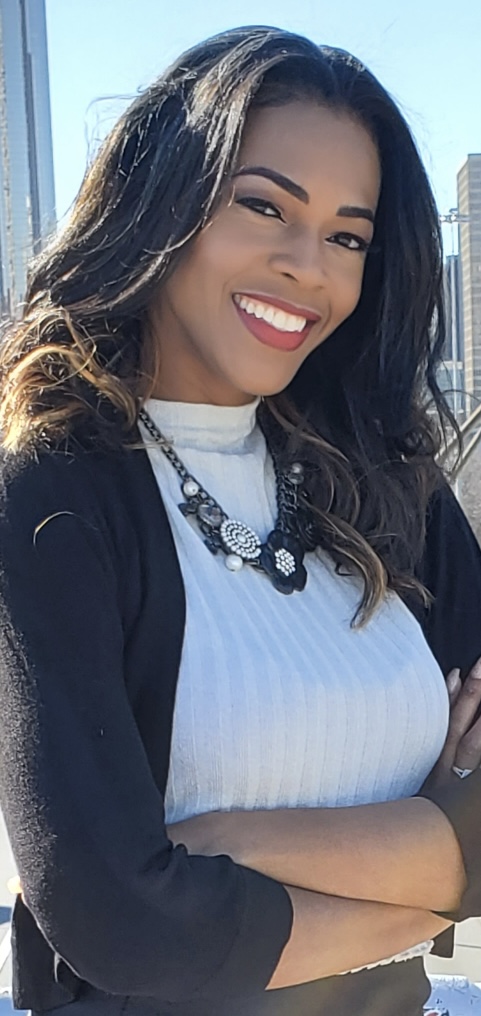 Smiling woman with long dark hair, wearing a white top and black cardigan, outdoors with city buildings in background.