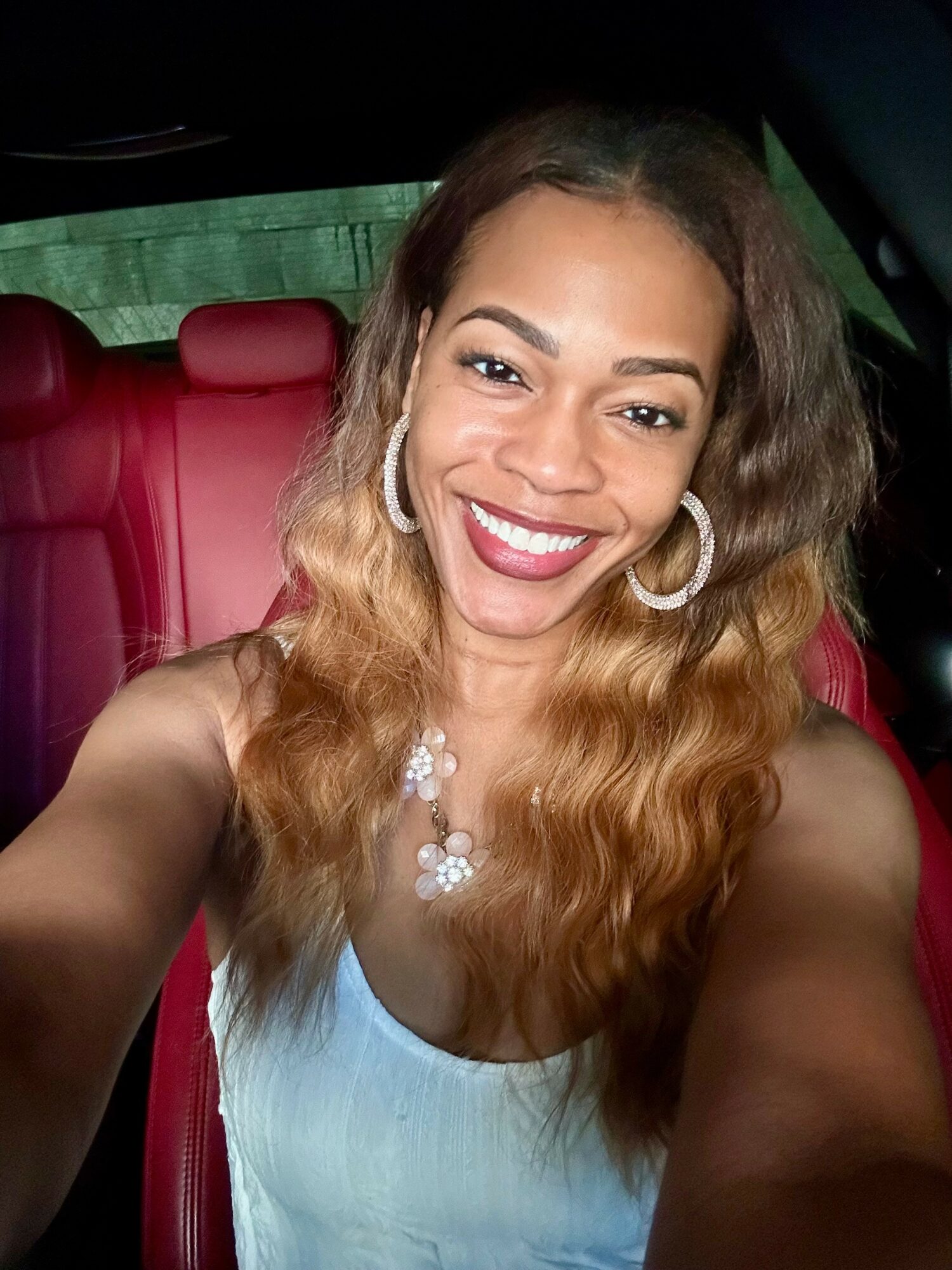 Smiling woman with wavy hair, wearing hoop earrings and a necklace, sitting in a car with red interior.