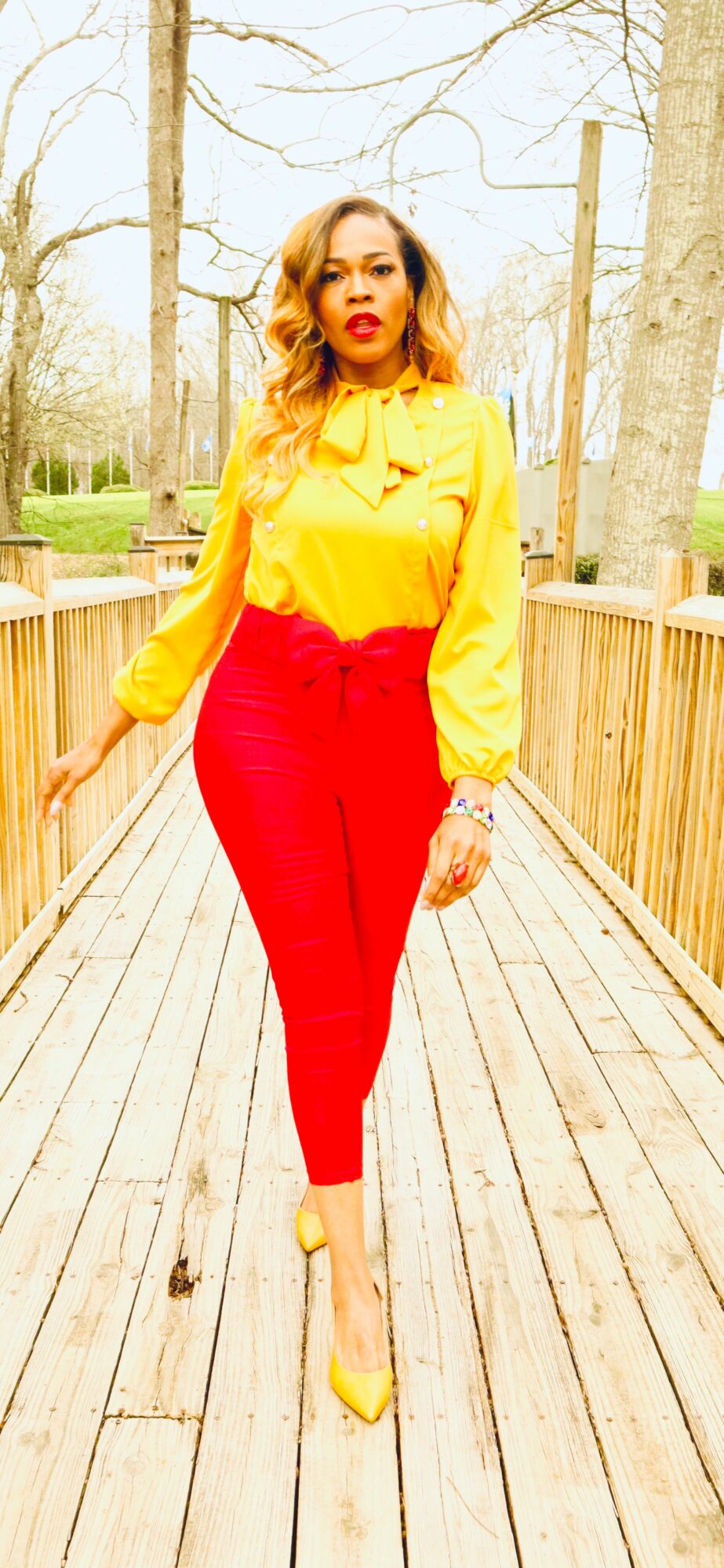 Woman walking on a wooden bridge in a park, wearing a yellow blouse and red pants, with trees in the background.