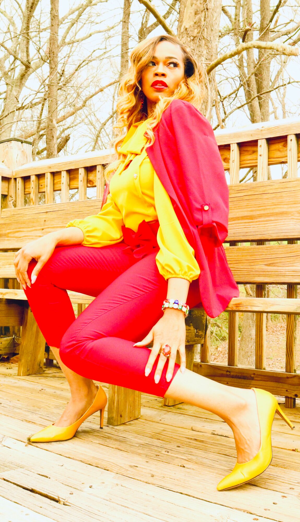 Woman in yellow and red outfit with yellow heels, sitting on a wooden bench outdoors, surrounded by trees.