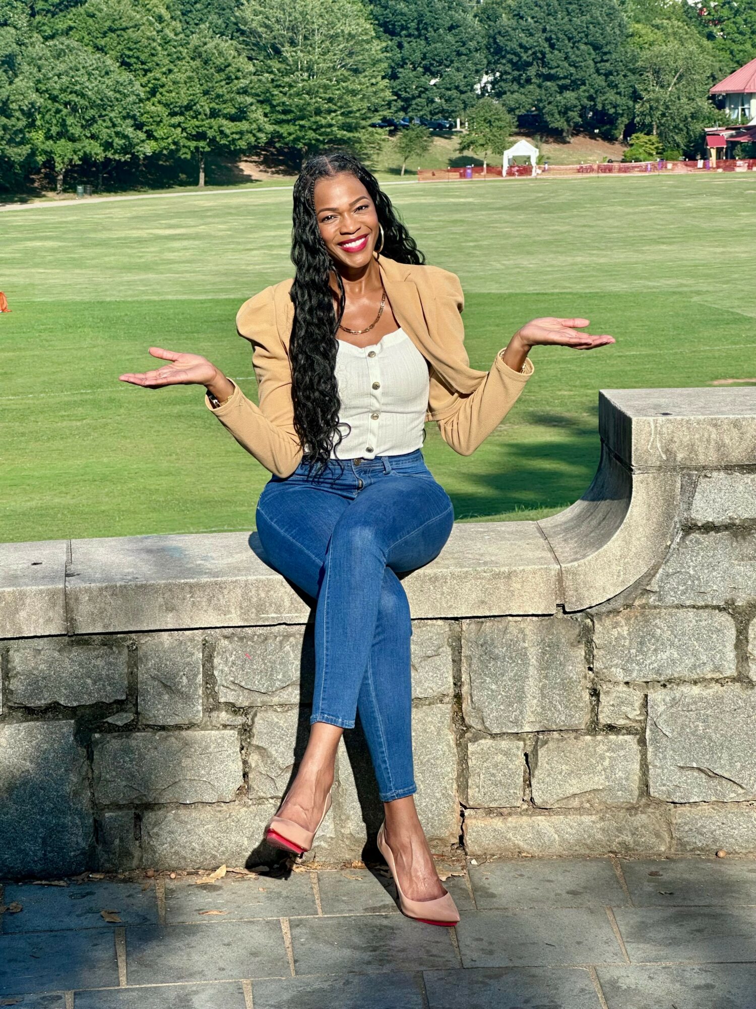Woman sitting on stone wall outdoors, smiling, with arms raised, wearing a beige blazer, white top, and blue jeans.