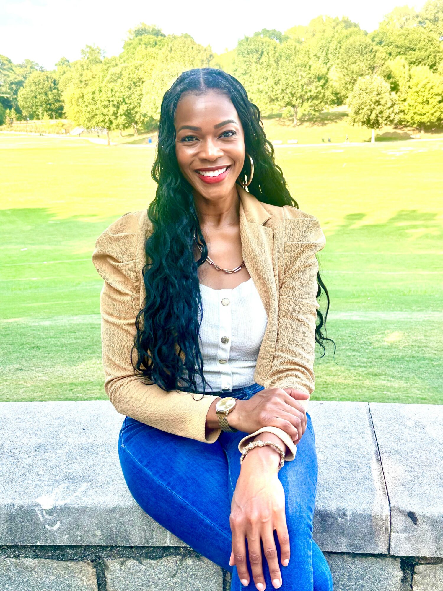 Smiling woman with long dark hair, wearing a beige blazer and white top, sitting outdoors on a stone ledge.