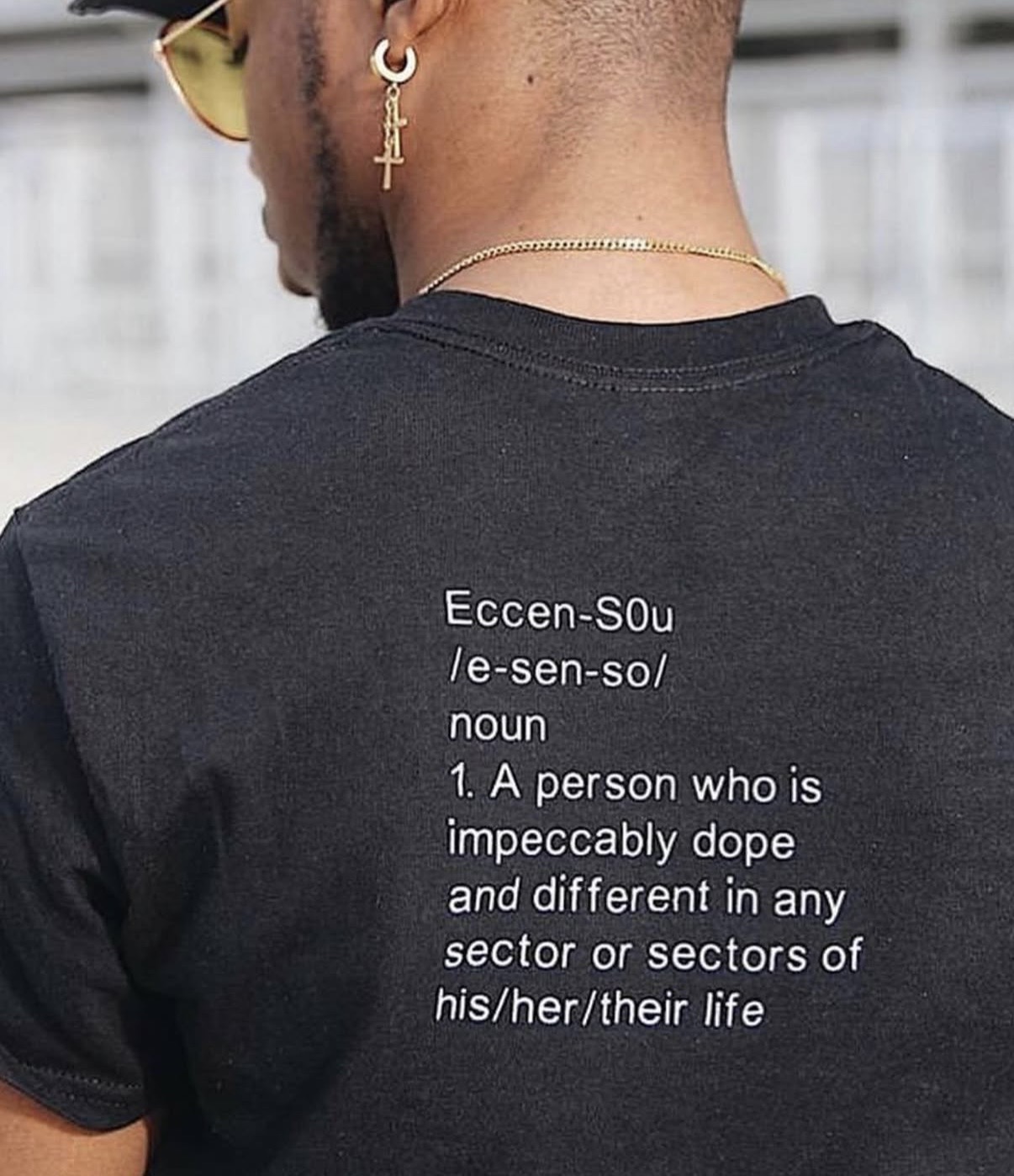 Back of a person wearing a black shirt with a definition printed on it, gold earrings, and a gold chain.