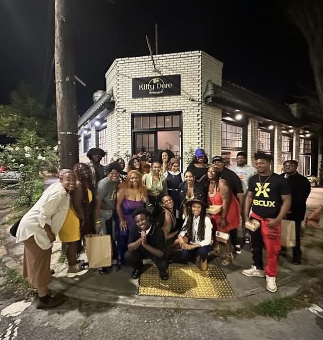 Group of people gathered outside a brick building at night, some holding shopping bags, smiling for the photo.