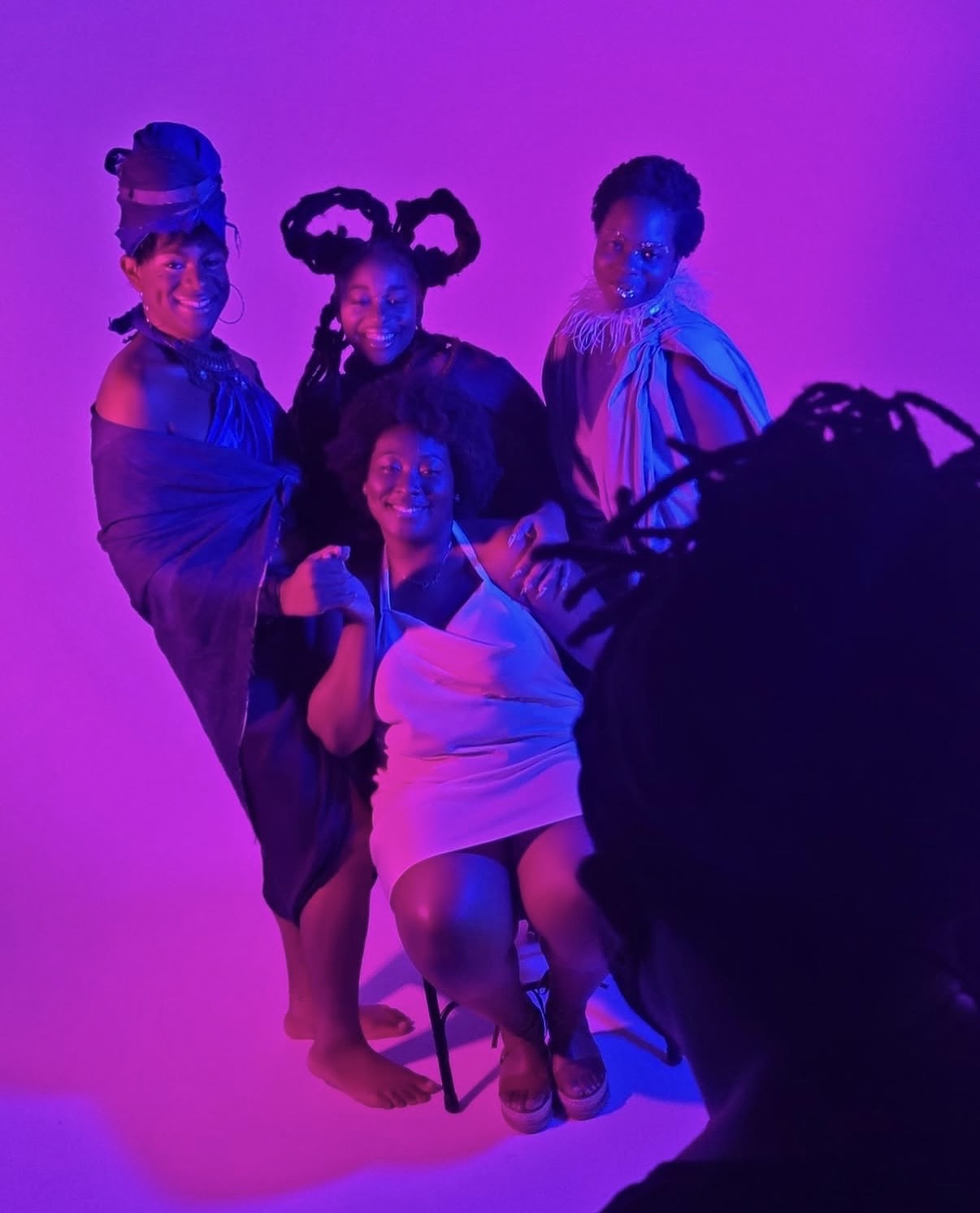 Four people pose against a purple background, with one seated and three standing, wearing colorful costumes and accessories.