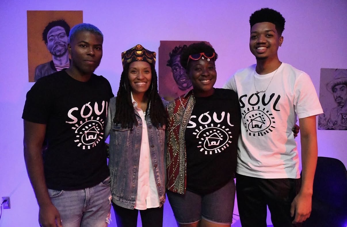 Four people standing together, smiling, in a room with purple lighting and artwork on the walls.