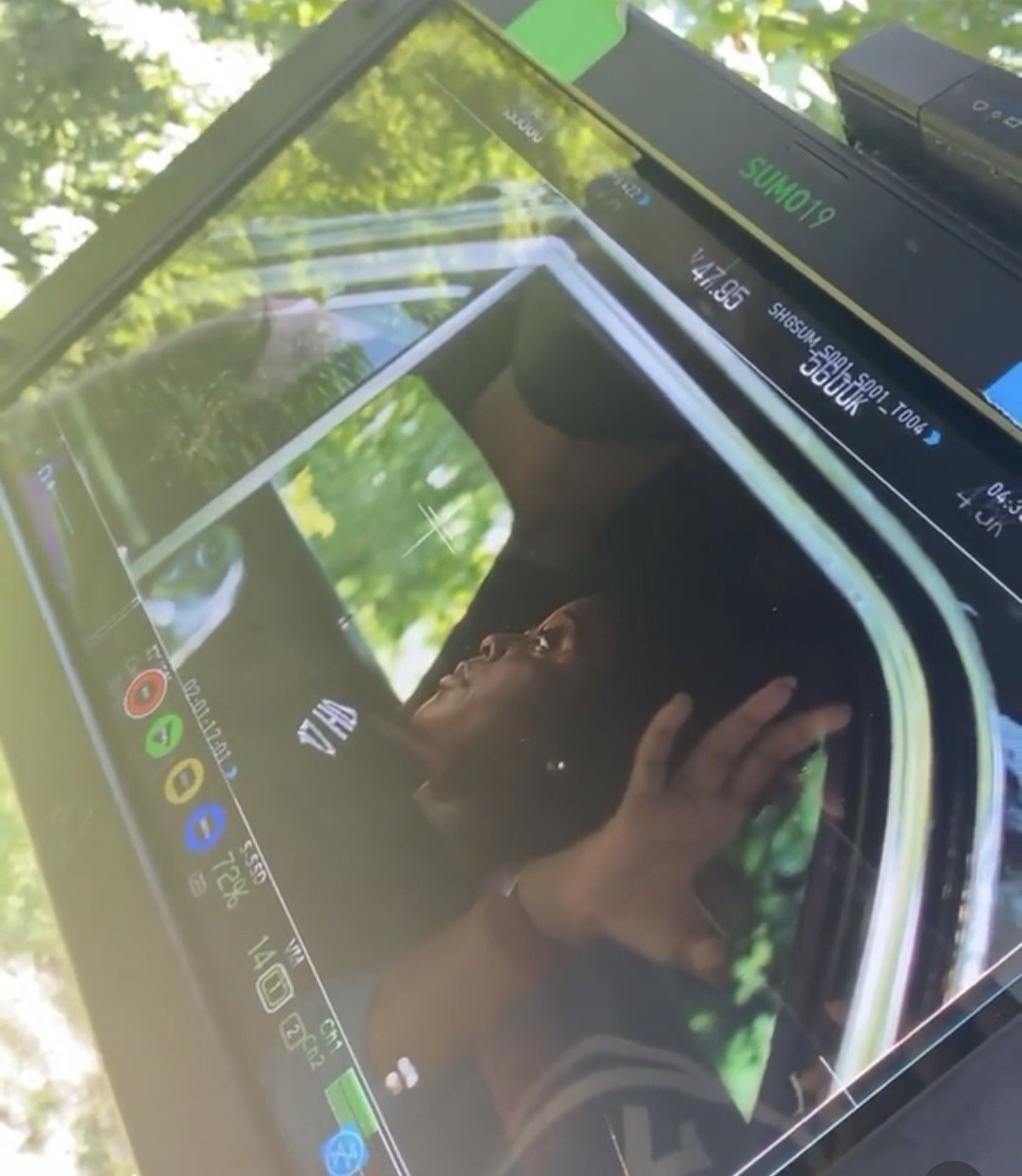 Person taking a selfie with a smartphone outdoors, surrounded by green trees and sunlight, reflected in a glass surface.