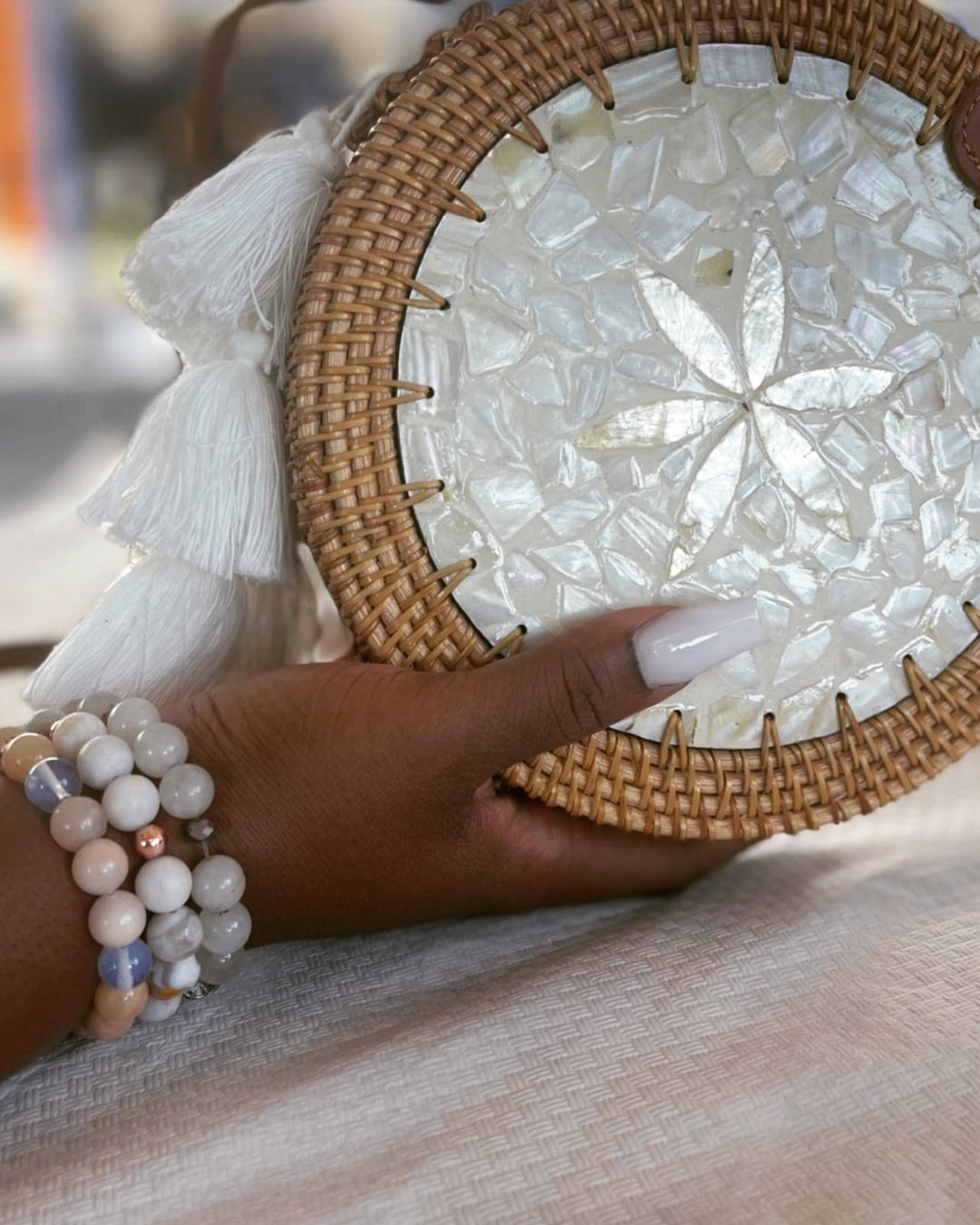 Person holding a round woven bag with a shell mosaic design, wearing a beaded bracelet, with a blurred background.