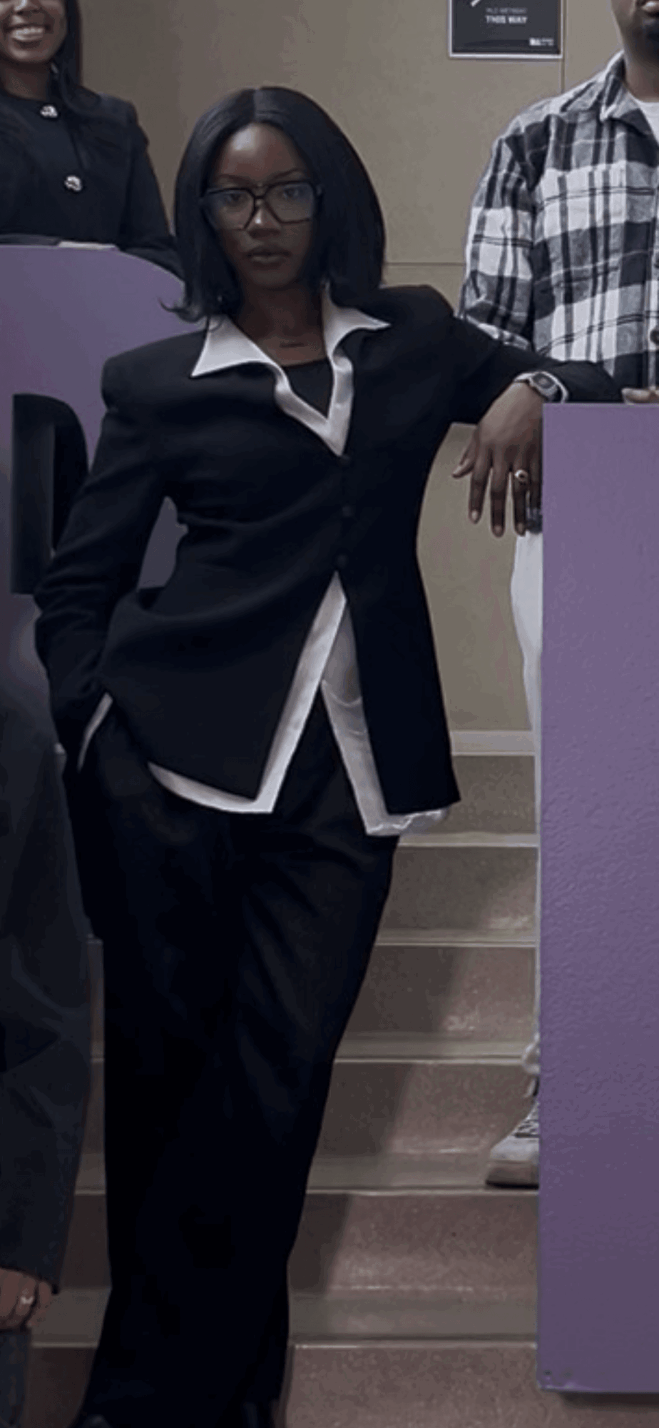 Woman with glasses in a black suit standing on stairs, with people around her, in an indoor setting.