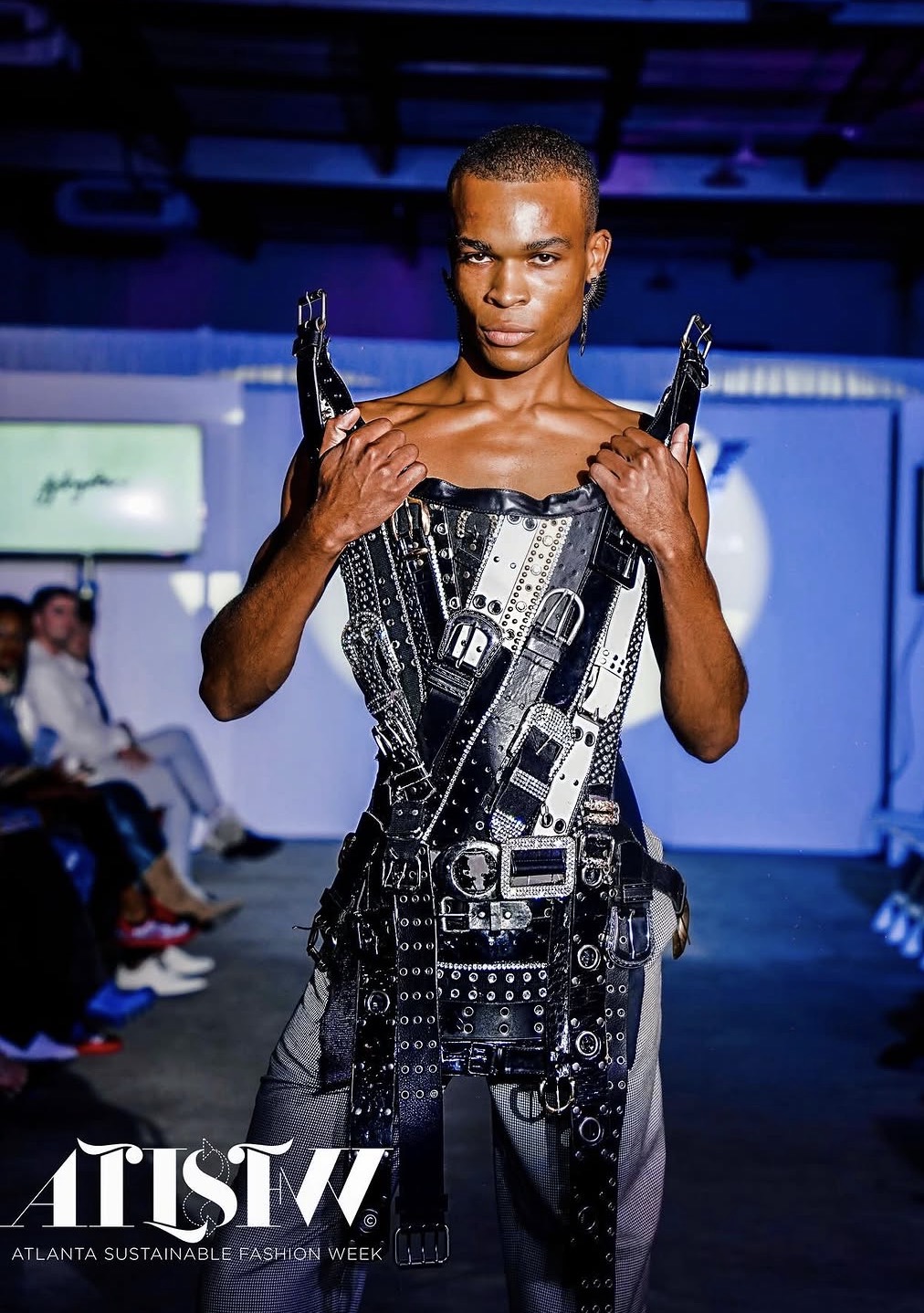 Model walking runway in black, metallic, harness-style outfit with straps and buckles, audience seated on sides, blue background, fashion show setting.