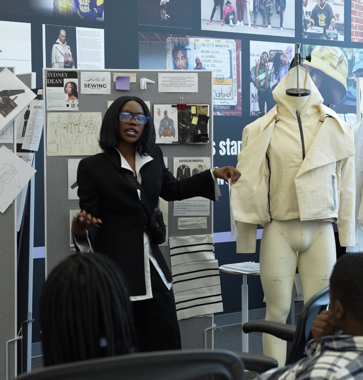 Woman speaking in front of a display with a mannequin dressed in a cream jacket, people listening, and a collage of images and text on the wall.