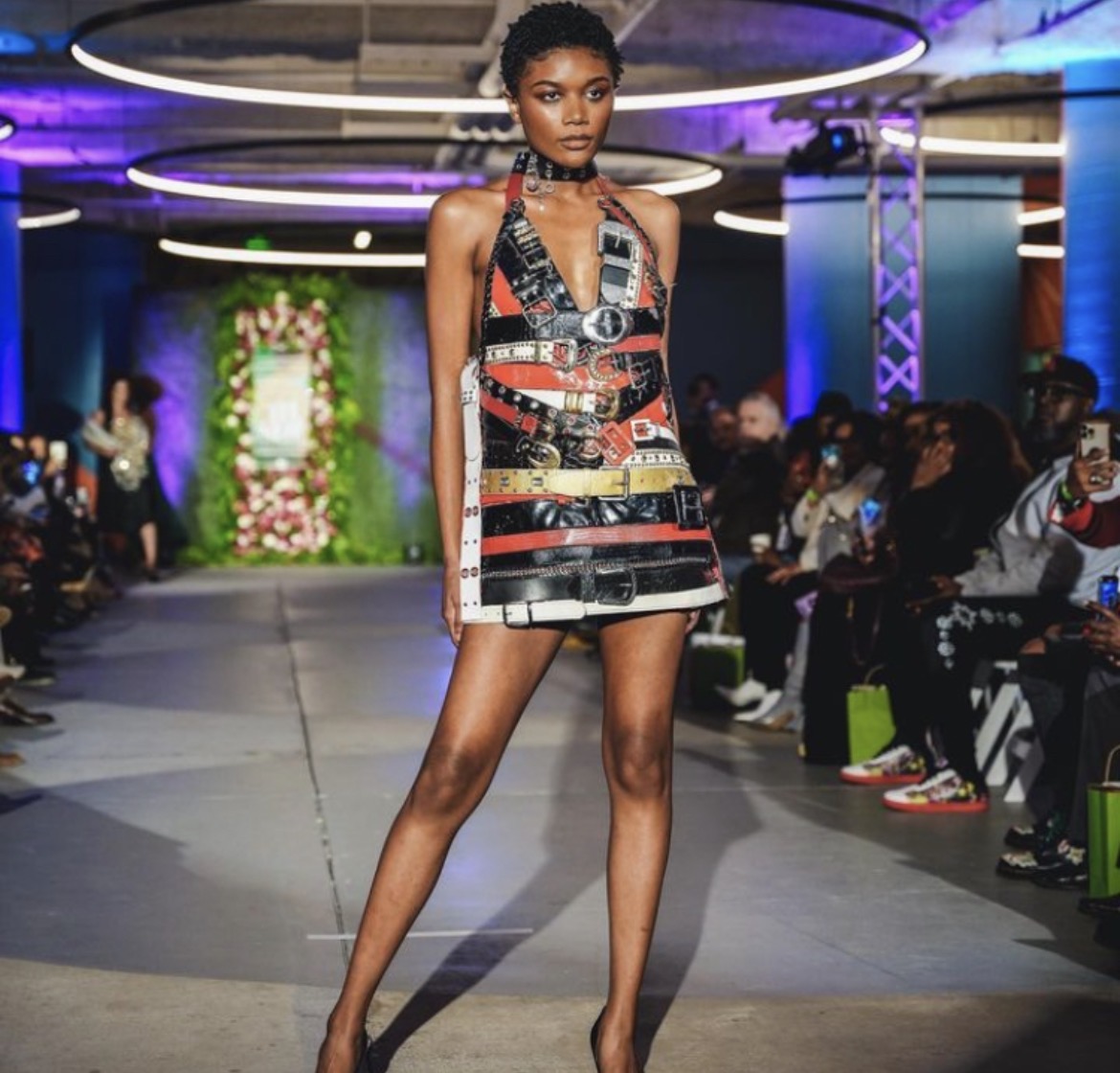Model walking on runway in a colorful dress with geometric patterns, audience seated on both sides, vibrant lighting overhead.