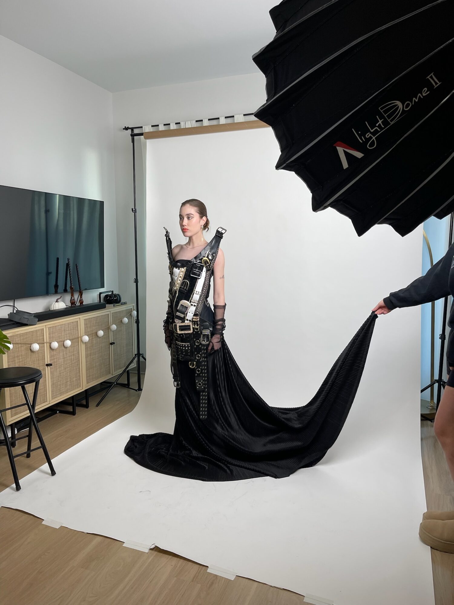 Young woman in black dress and harness stands in front of a white backdrop during a photo shoot, with a large black umbrella overhead.