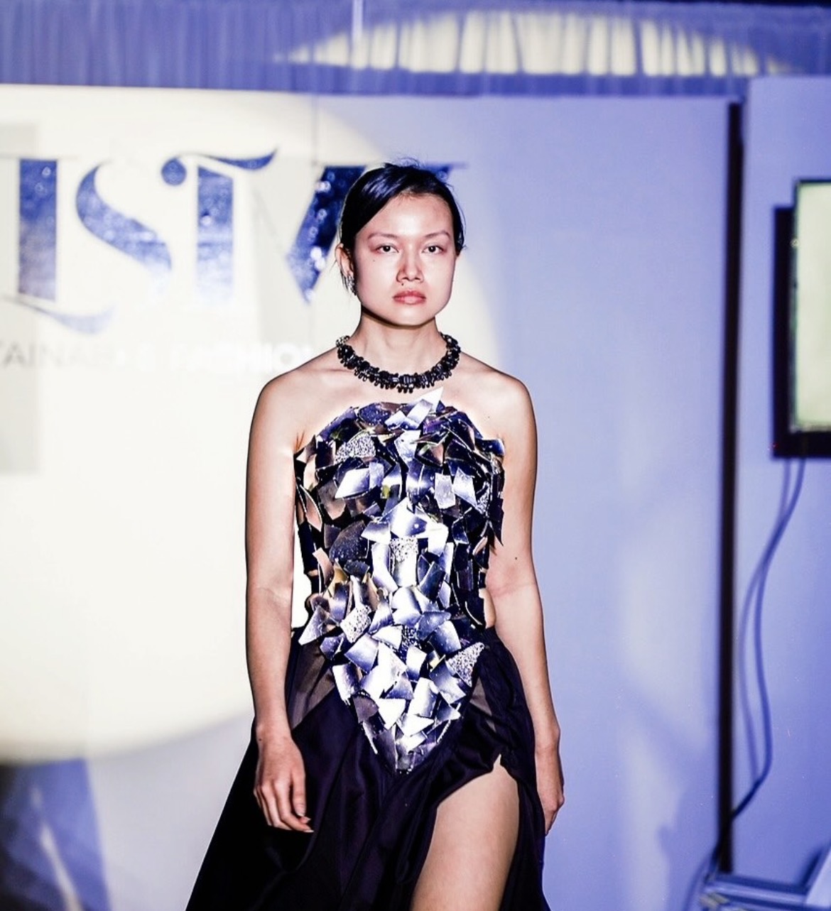 Young woman in a strapless dress with a metallic pattern, standing indoors with a blue and white background.