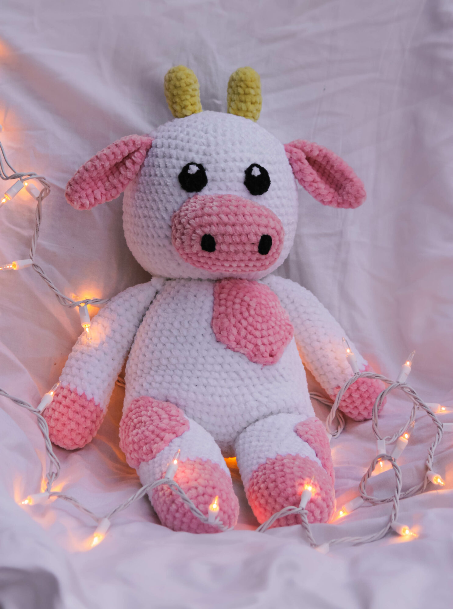Knitted plush cow with pink spots, black eyes, and yellow horns, sitting on a white fabric with fairy lights.