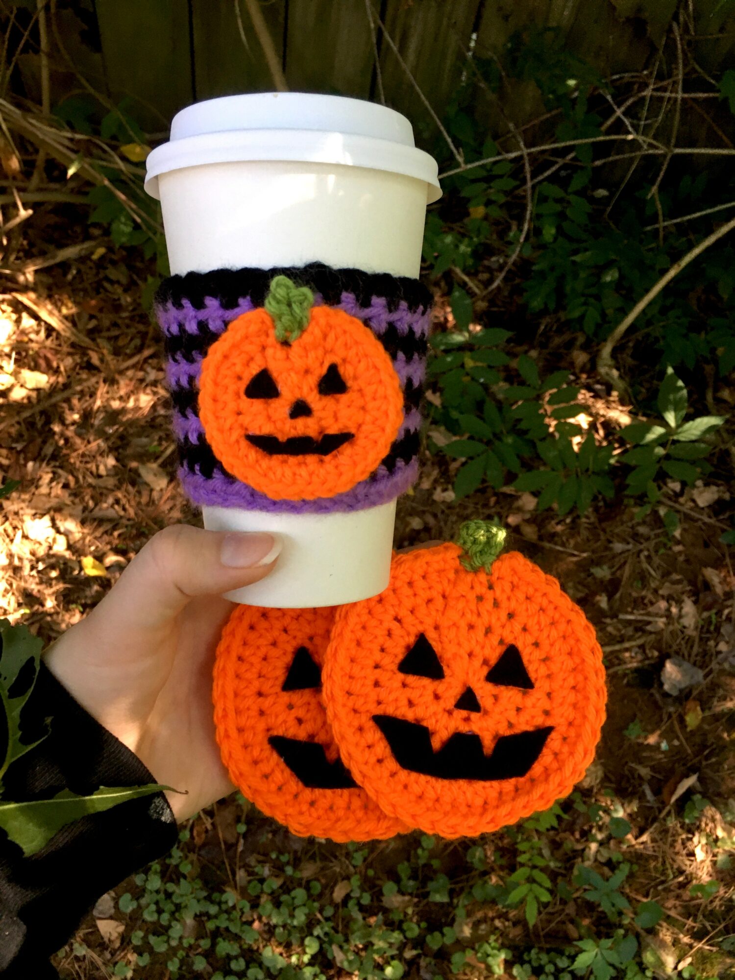 Hand holding a coffee cup with a purple and black knitted sleeve featuring a pumpkin decoration, and a larger knitted pumpkin decoration below, outdoors.