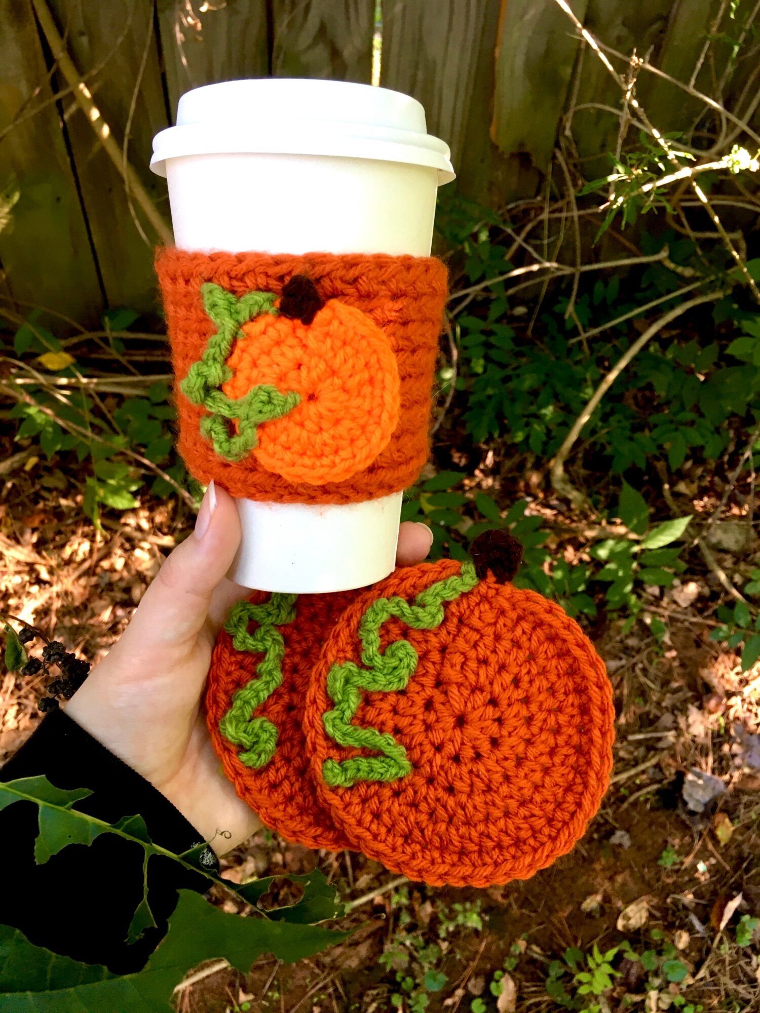 Hand holding a coffee cup with a pumpkin and vine crochet cozy, surrounded by outdoor plants and leaves.